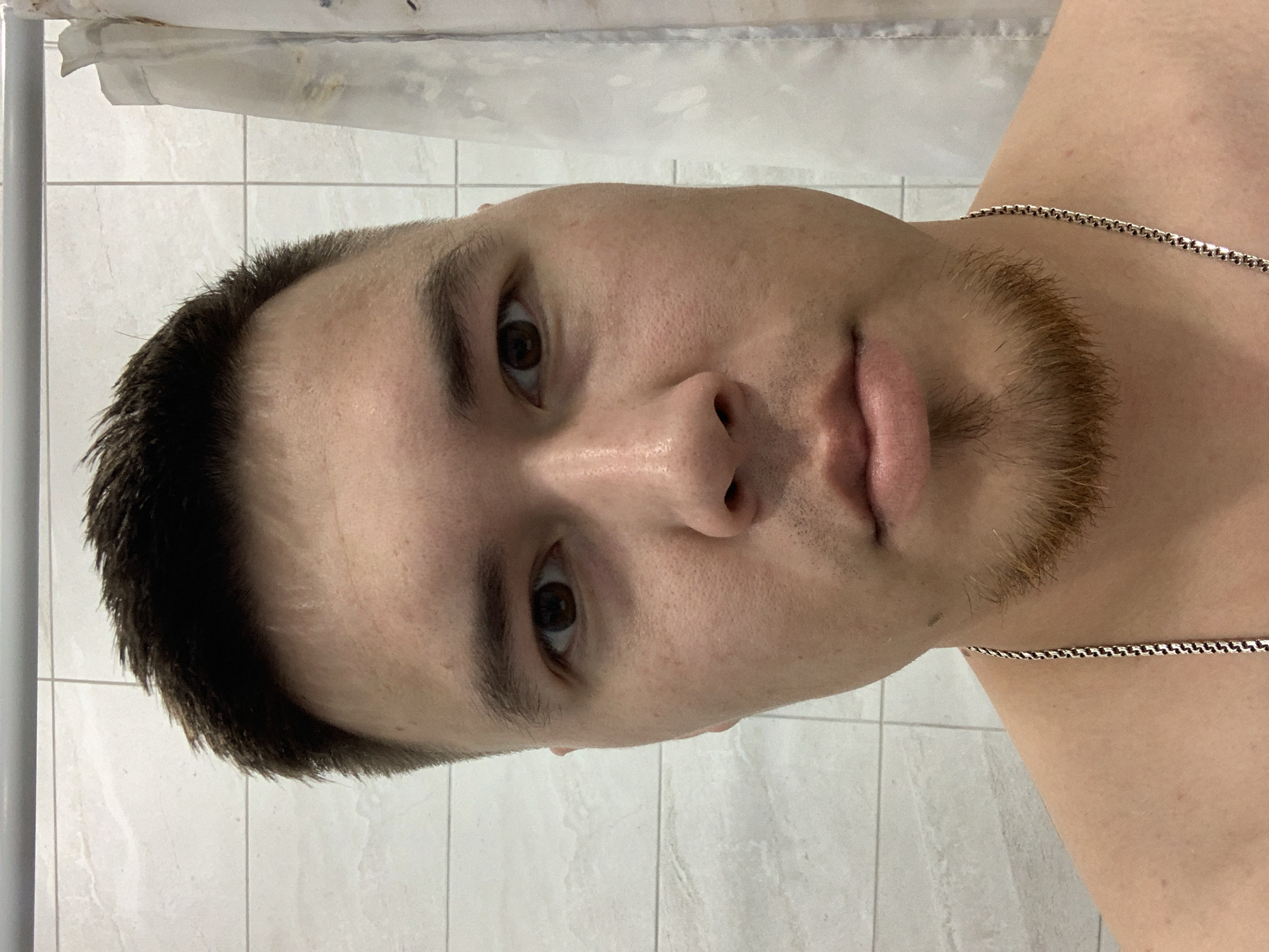 A close-up, eye-level, forward-facing portrait of a young adult male shows him from the shoulders up. He has short, dark hair, styled upward and to the front, with a short, light brown goatee. His eyes are dark brown. He is wearing a silver chain necklace. The background consists of white tiled walls and a white, patterned shower curtain. The image appears to have been taken in a bathroom, possibly in Stavropol, Russia, based on the available information.