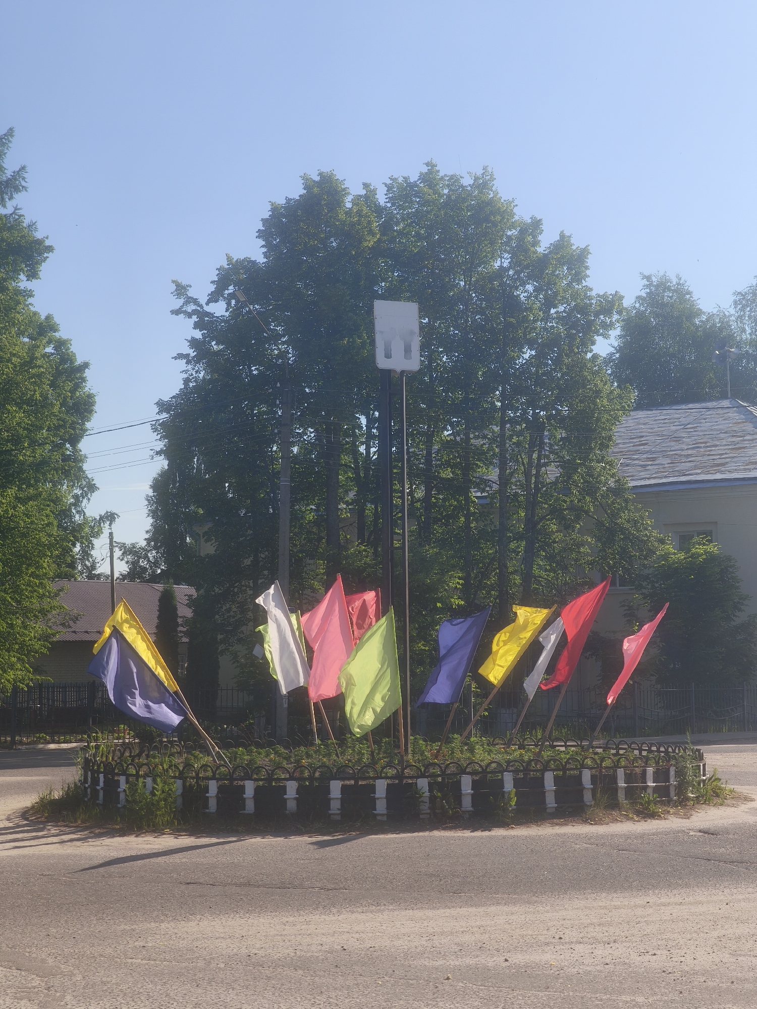 The image captures a view of a roundabout in Kady, Russia, adorned with a circular arrangement of brightly colored flags. The flags, predominantly in hues of yellow, blue, pink, and red, are attached to poles and are fluttering in the breeze. In the center of the roundabout, a tall pole stands topped with a white, rectangular structure, possibly an antenna or a light fixture. The roundabout is surrounded by a decorative black and white metal fence. Beyond the roundabout, lush green trees form a backdrop, and a building with a grey roof is visible in the distance. The ground is paved with asphalt, showing some wear and tear. The sky above is clear and blue, suggesting a sunny day. There are no people or vehicles present in the immediate vicinity of the roundabout. The overall scene depicts a public space decorated for an event or as a permanent feature of the town's landscape.