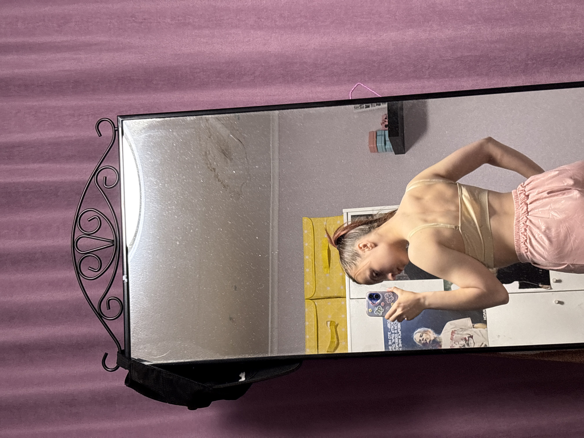A mirror reflects a young woman taking a selfie with a blue smartphone in what appears to be an indoor setting, potentially a bedroom or dressing area. The mirror's frame is ornate with decorative curls. The background includes a mauve-colored wall, part of a yellow storage unit, and a white cabinet with a poster attached to the side. The woman is wearing a beige top and pink shorts. The reflection also shows other items in the room, such as books stacked on a shelf and a black object hanging from the mirror. There is no indication of the city of Прикамский, Russia in the image.