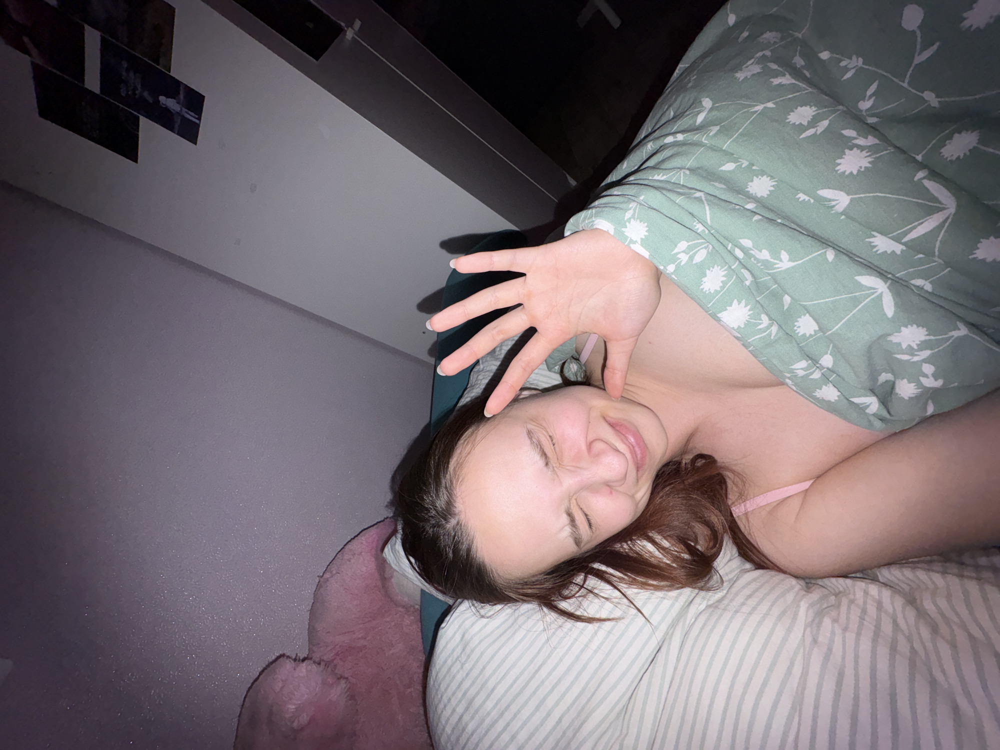 A young woman lies in bed with a light green floral patterned blanket partially covering her body. Her hand is raised in front of her face, as if shielding her eyes from light. She is smiling with her eyes closed and head turned slightly to the left. The pillow is striped, and a pink stuffed animal sits behind her head against the wall. The wall is light gray, and the room is dimly lit. Behind her head, there's a white wardrobe or closet. A few framed photos hang on the wall near the wardrobe. The location is indicated as Прикамский, Russia.