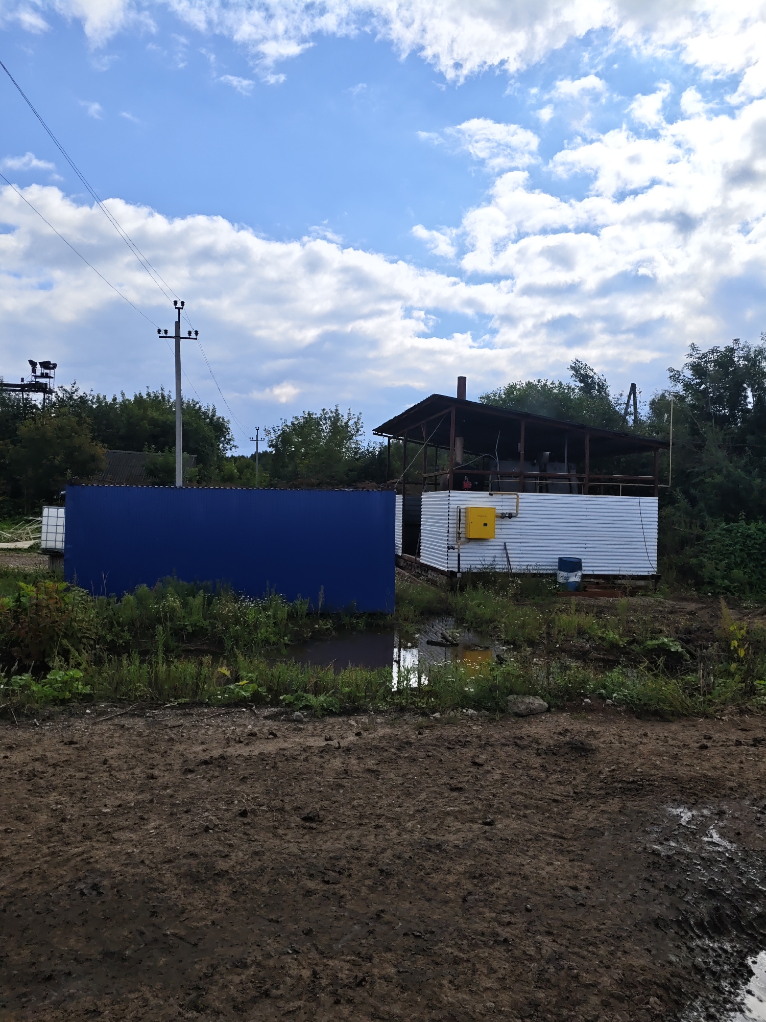 The image depicts an outdoor scene in the village of Platoschino, Russia. In the foreground, a muddy, dirt track with puddles suggests recent rain. Behind the track, a tall, bright blue corrugated metal wall stands prominently, bordered by overgrown grass and wildflowers. To the right of the blue wall, a white corrugated metal building with a dark-colored, open-sided roof is visible. This structure appears to be an industrial or utility building, with a yellow gas meter box mounted on its exterior wall and a blue barrel placed on the ground nearby. A metal chimney protrudes from the roof of this building. Trees and dense foliage surround the structures, and power lines and utility poles are visible in the background against a sky filled with white, puffy clouds. The overall scene suggests a rural or semi-industrial setting.
