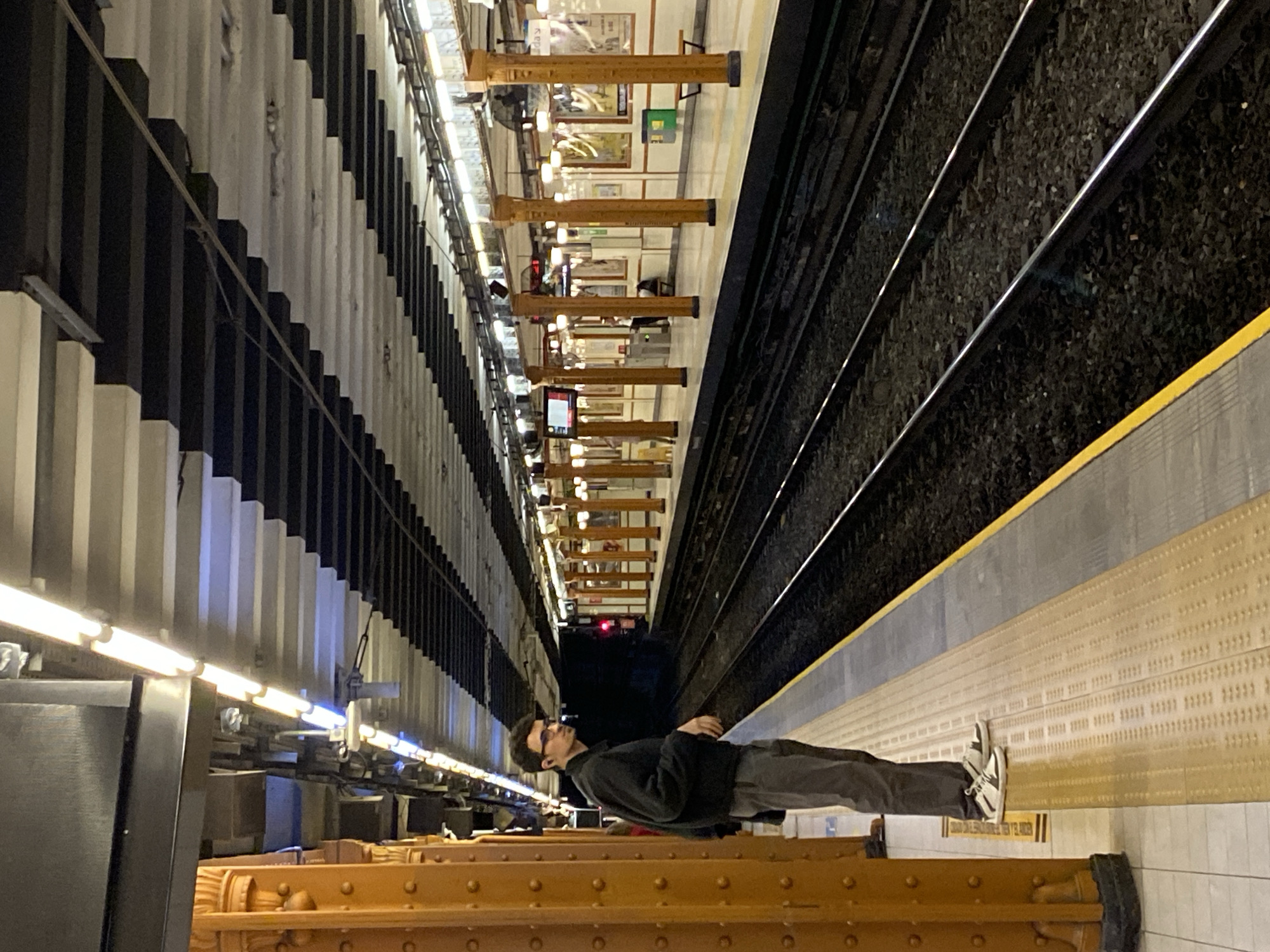 The image depicts a man lying on a train platform in the Buenos Aires, Argentina subway system. The platform has a yellow safety strip and a textured surface. The man is wearing glasses, a dark-colored jacket, and pants, and white sneakers. His head is oriented towards the tracks, and his body is parallel to the edge of the platform. The subway tracks are visible next to the platform edge. The opposite side of the subway station has dark and light striped walls with rows of lights, and ornate columns. The far end of the subway tunnel is dark, with a visible red light.