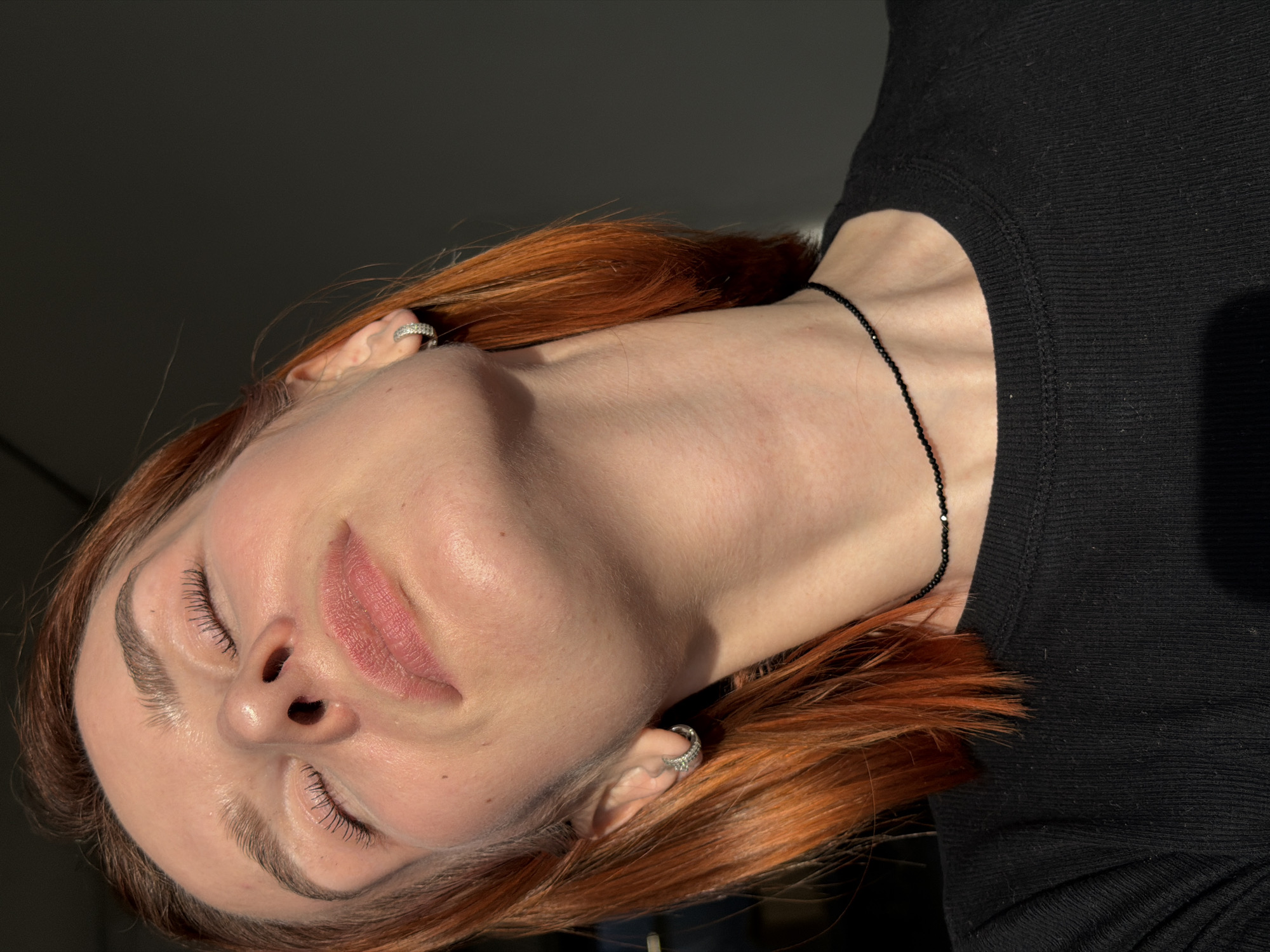 A medium shot of a light-skinned woman with red hair wearing a black top and a black beaded necklace. She is facing upwards with her eyes closed. The woman has silver earrings in both ears. The background is a plain, dark color. The location is not discernible.