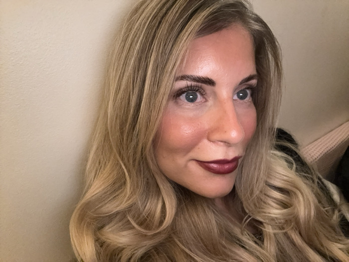 A blonde woman with wavy hair looks to her right. Her complexion is fair, and she is wearing makeup, including dark lipstick. She has light-blue eyes and dark eyebrows. The background is a plain wall, and a glimpse of a pink blanket is visible to the right. The lighting appears to be coming from the left side of the frame.