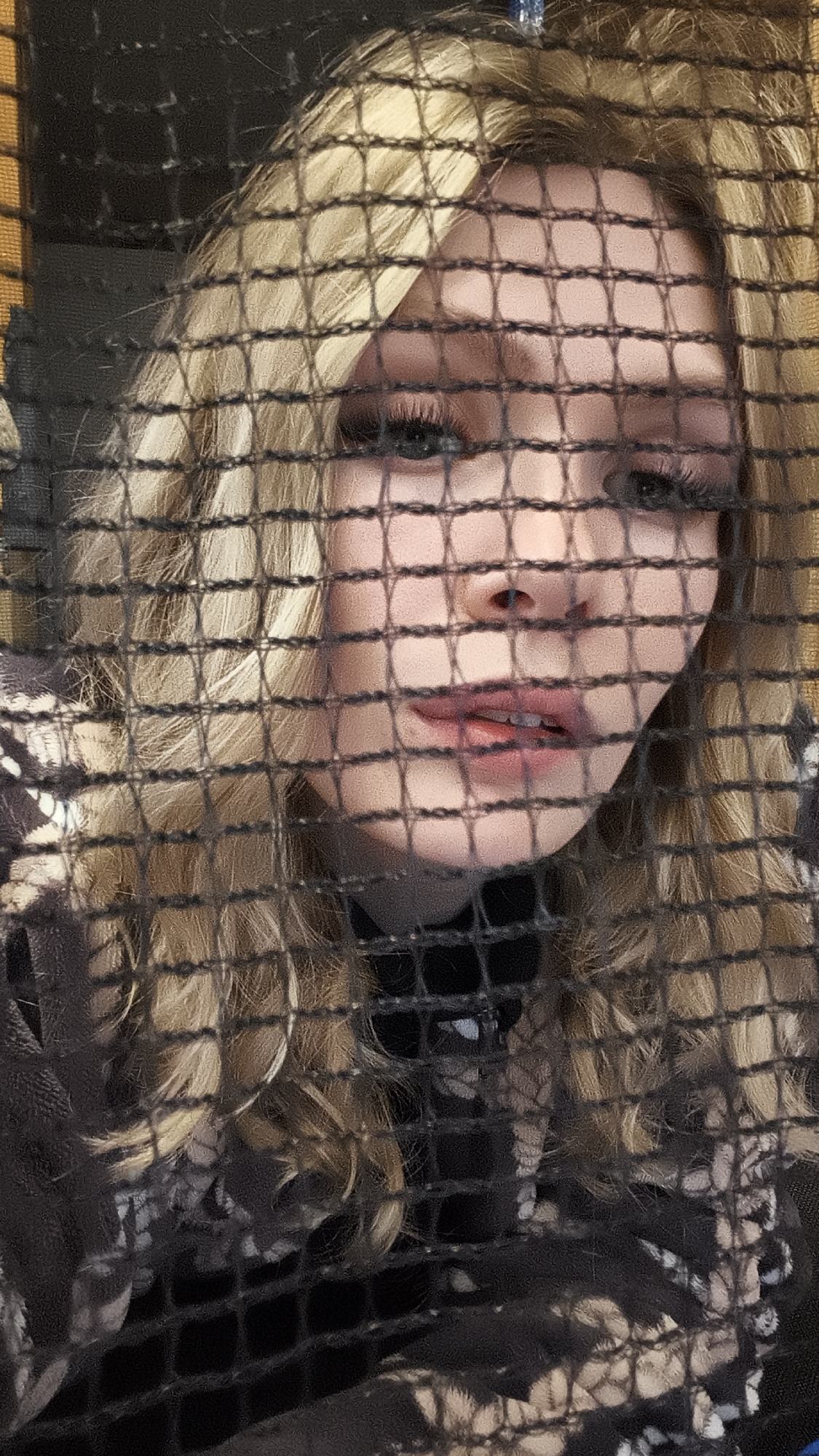A person with blonde hair is positioned behind a black net. The individual's face is partially visible through the netting, with details like eyes, nose, and mouth discernible. The person is wearing a dark-colored turtleneck sweater or garment with a patterned design visible beneath. The environment appears to be indoors. Location information, Русский Вожой, Russia, is not relevant to the visual details of the image.