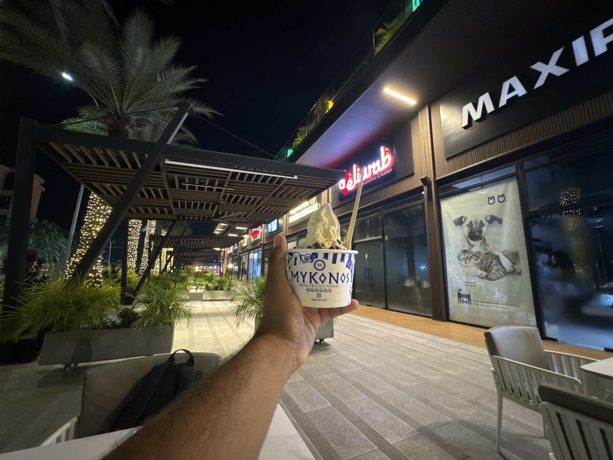 A first-person perspective photo captures a person holding a cup of "Mykonos Greek Frozen Yogurt" at night. The setting appears to be a modern outdoor shopping area. The person's arm and part of their torso are visible in the foreground. A backpack rests on the seat next to them. Beyond the person, there are planters with greenery, decorative lights on trees, and covered walkways.
In the background, several storefronts are visible, including one with a sign that reads "MAXIF" and another with an Arabic inscription. A poster of a dog and cat is visible. The flooring is paved with gray tiles. The picture seems to have been taken at a table setting. The city is Pampatar, Venezuela.
