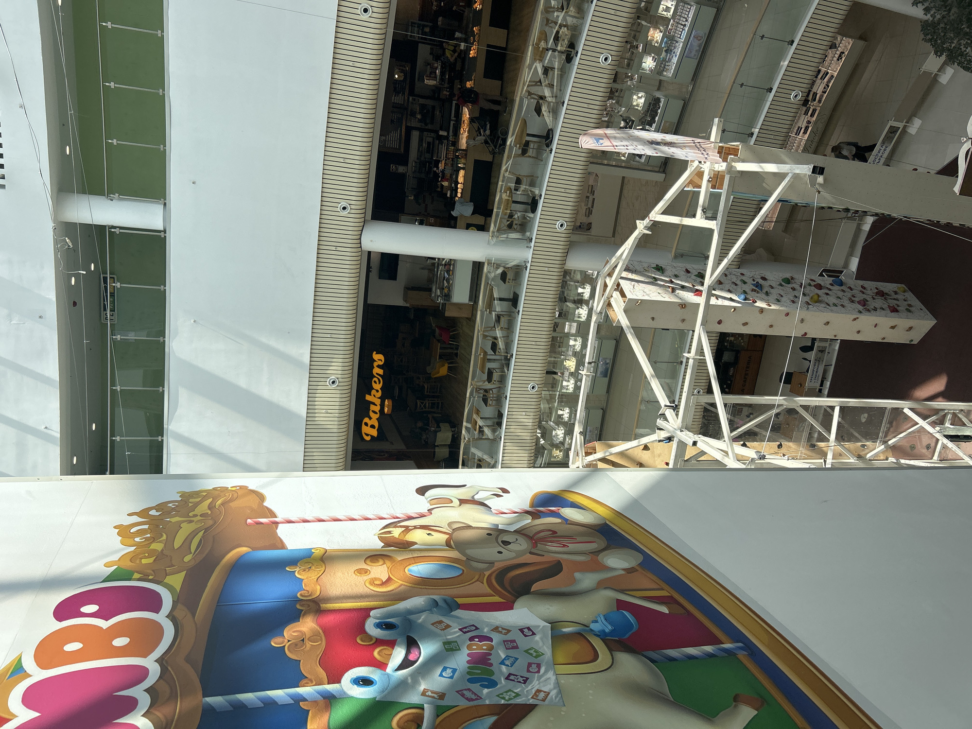 The image shows the interior of a shopping mall in Plovdiv, Bulgaria, viewed from an elevated position. The foreground features a section of a colorful carousel mural with cartoon animals. In the background, several levels of the mall are visible, including shops and dining areas. A "Bakers" sign can be seen on one of the floors. There is a rock climbing wall structure. The lighting appears to be artificial, coming from the mall's fixtures. The photograph is taken at an angle.