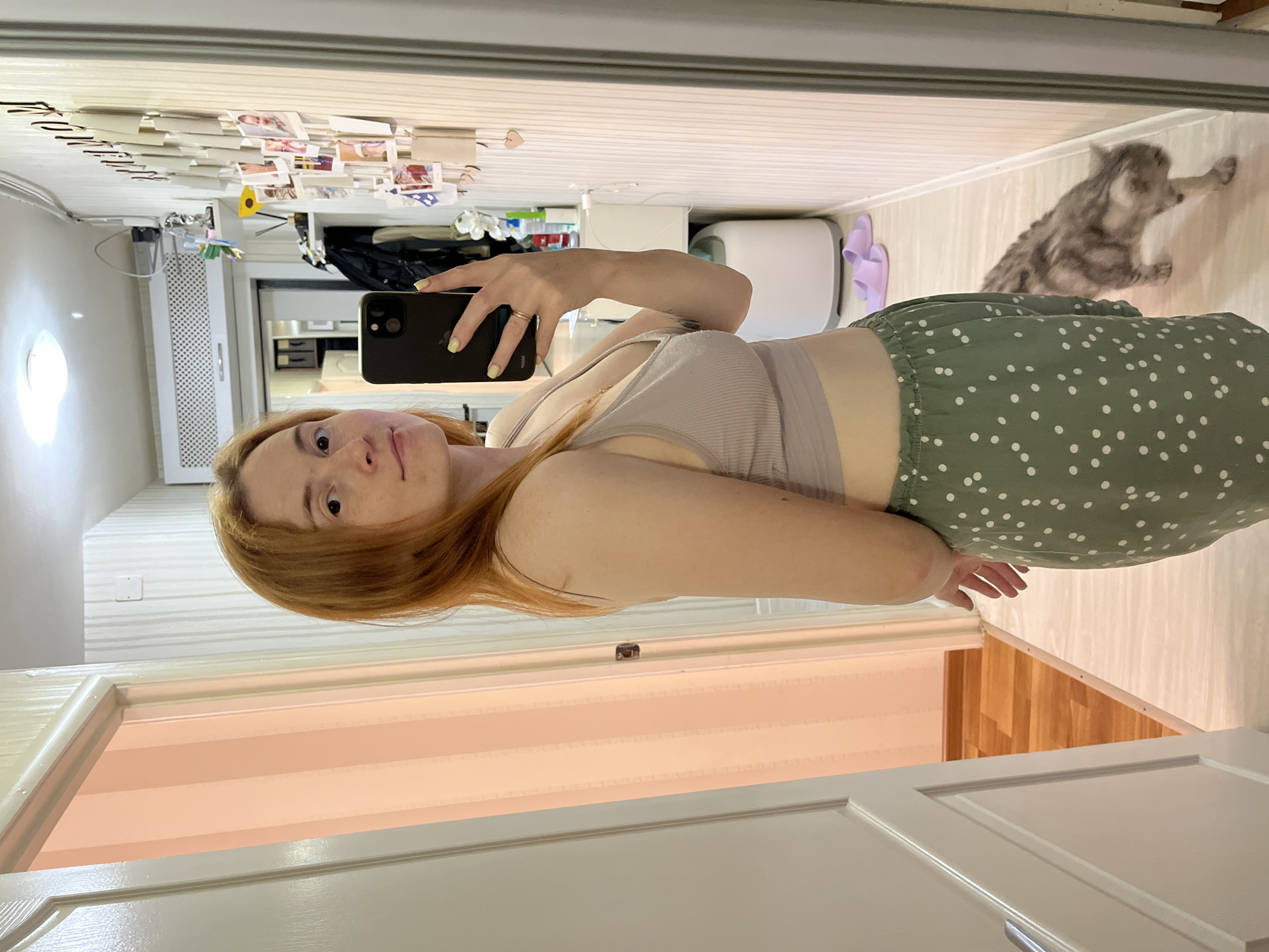A young woman captures a mirror selfie in what appears to be a home interior in Chaikovsky, Russia. She has reddish-brown hair and is wearing a light grey ribbed crop top and a green skirt with white polka dots. Her left hand holds a dark-colored smartphone, showing light yellow nail polish and a ring on her finger. She looks directly into the camera with a neutral expression.
The mirrored reflection reveals a domestic scene. To the woman's right, a grey tabby cat is seen on a light wooden or laminate floor, stretching or moving. Nearby, a pair of purple flip-flops rests on the floor. The background features a white wall with horizontal textured stripes, a rectangular white vent, and a circular light fixture. Above the vent, a shelf is cluttered with various small items, including numerous photos or cards pinned to it, and several dark hair clips. The overall setting suggests a bedroom or dressing area. Part of a white paneled door is visible in the foreground outside the mirror's reflection. The lighting is bright, consistent with indoor artificial light or a well-lit room during the day. No discernible text is visible in the image.