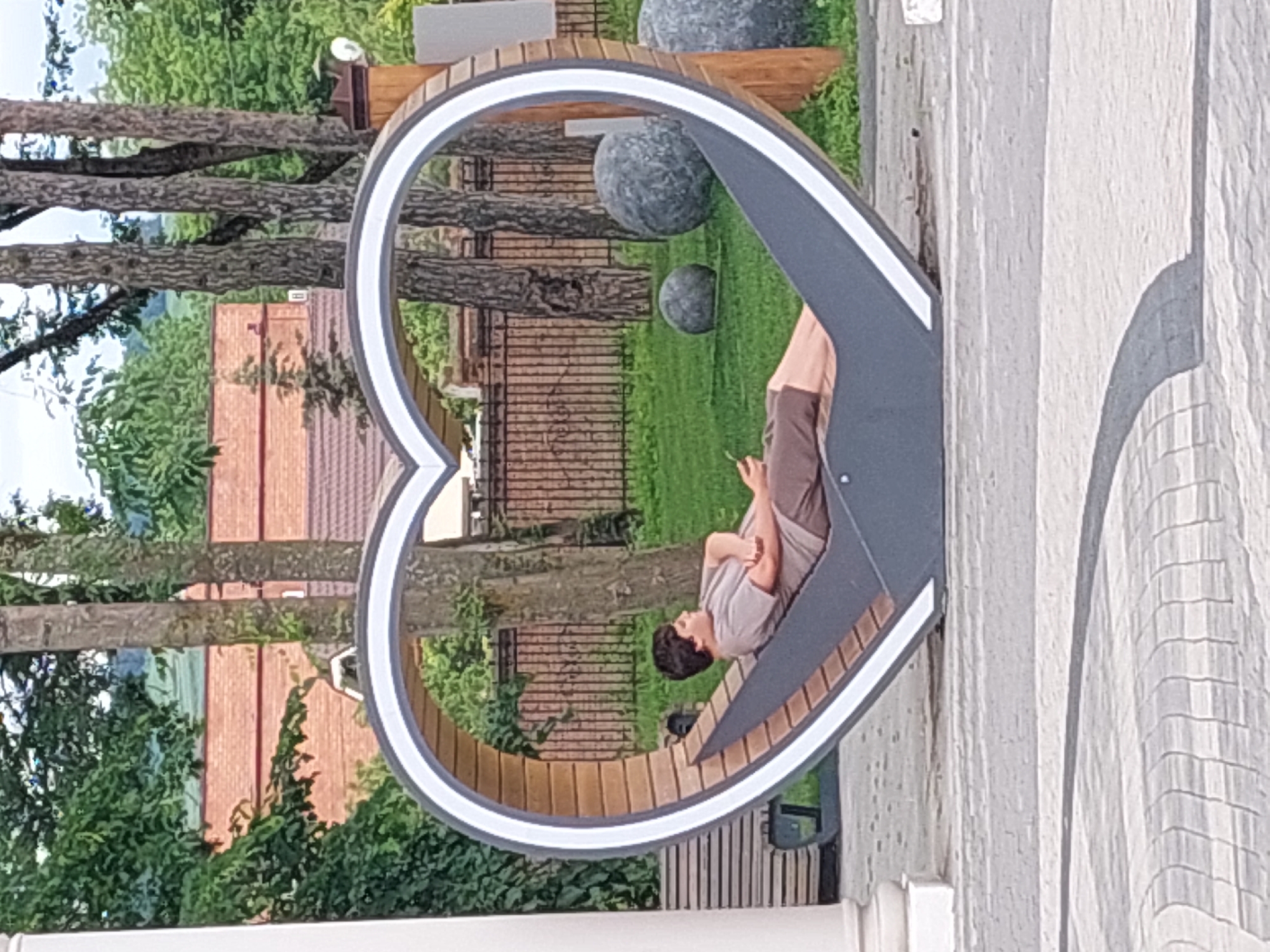 A person is reclined on a heart-shaped bench in what appears to be a park in Alikonovka, Russia. The bench is made of wood and metal, with a white trim around the heart shape. The person, dressed in a t-shirt and shorts, is holding a phone in their hand. In the background, there are trees, a brick building, a fence, and large decorative spheres. The ground is paved with cobblestones.
