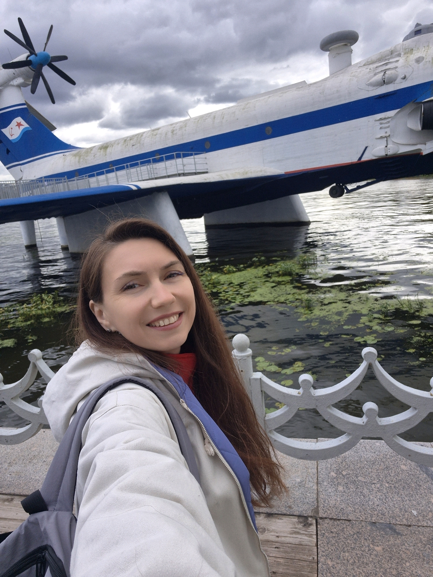 A young Caucasian woman with long, brown hair and a friendly smile takes a selfie outdoors. She is wearing a light-colored hooded jacket over a red top and a blue vest. A gray backpack is slung over her left shoulder. Behind her, a large, white seaplane with blue stripes is docked in dark water, which is covered in patches of green aquatic plants. A white decorative fence is in the foreground. The sky above is overcast and cloudy. The seaplane appears to be a monument, possibly located at Gorky Park on the Moskva River in Moscow, Russia, a common location for such exhibits.