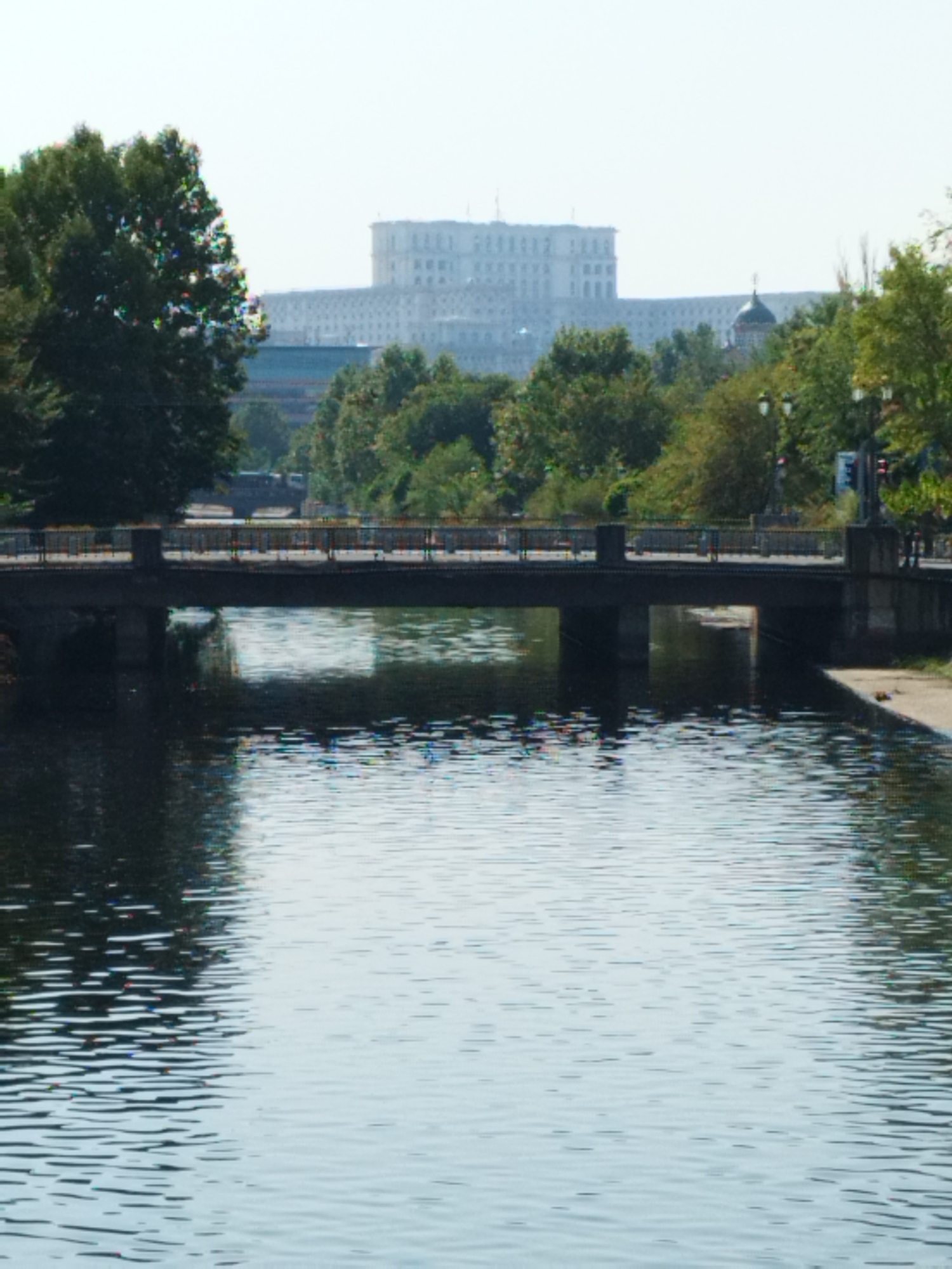 The image depicts a wide river or canal with a bridge spanning across it. The bridge is made of concrete and has railings along its sides. On the far bank, behind a line of trees, is a large, imposing white building with many windows, characteristic of the Palace of the Parliament in Bucharest, Romania. The sky is overcast and hazy. The water is calm, reflecting the bridge and the trees on the banks. There are no people visible in the image. The overall scene suggests an urban landscape with natural elements.
