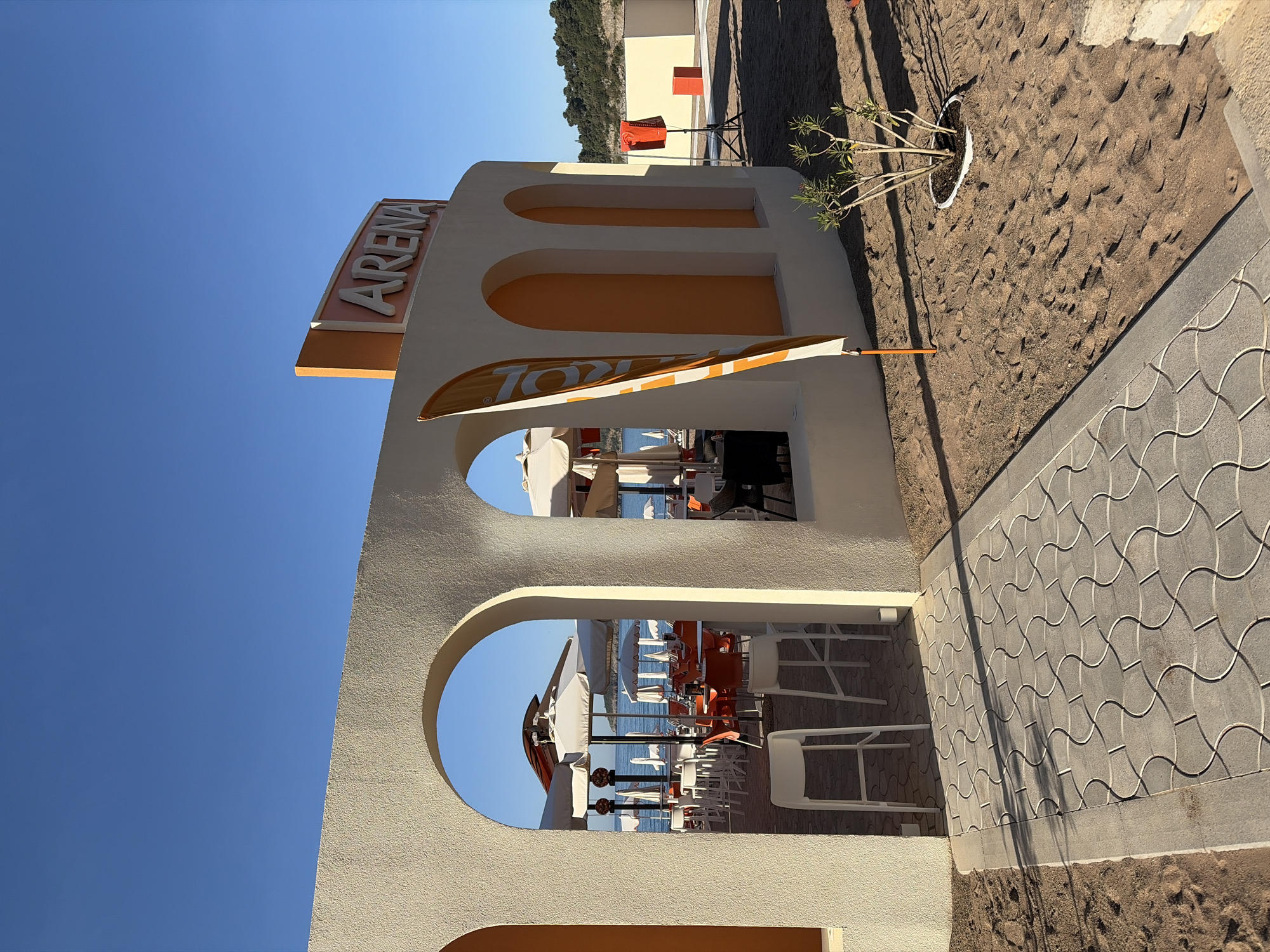 The image shows the "Arena" beach bar in Boreti, Montenegro, taken from a low angle looking up. The structure is a white building with orange accents, featuring arched openings that frame views of the Adriatic Sea and outdoor seating areas with white chairs and tables under beach umbrellas. A sign displaying "Arena" is visible at the top of the building. A sandy beach surrounds the bar, with a tiled walkway alongside the structure. A small potted plant sits on the sand near the building. The sky is clear and blue. The overall atmosphere is bright and sunny.