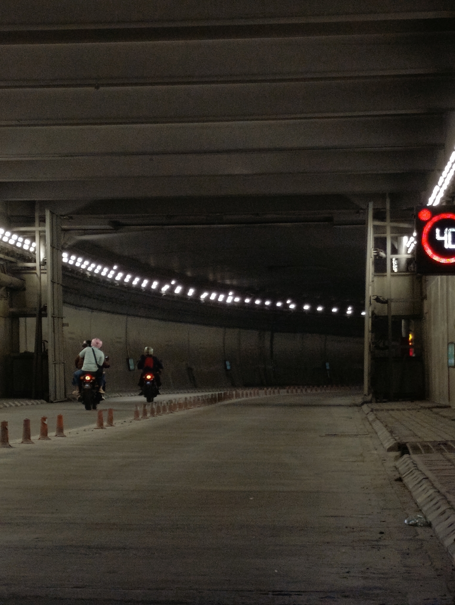The image shows a view inside a concrete tunnel. Two motorcycles, each with a rider, are traveling away from the viewer. The motorcycle on the left is ridden by a person wearing a light blue shirt and a pink helmet. The motorcycle on the right is ridden by a person wearing a dark colored jacket. The tunnel is lit by rows of bright lights along the ceiling. Orange traffic cones line the sides of the road. A digital speed limit sign displaying "40" is visible on the right side of the tunnel. The overall environment appears to be a roadway or passage within an urban infrastructure.