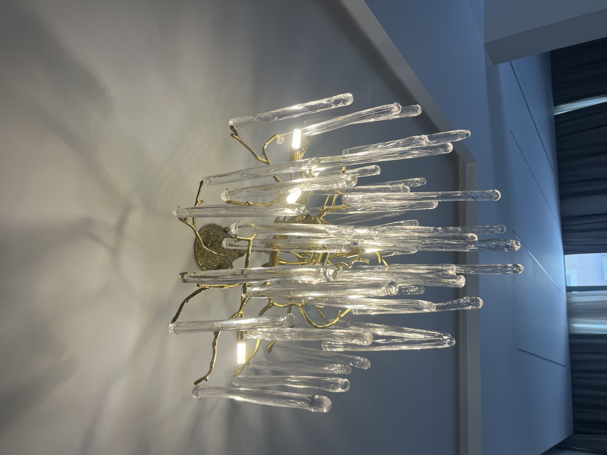 The image displays a close-up, low-angle shot of a modern chandelier hanging from a white ceiling. The chandelier features a gold-colored, branch-like structure from which numerous clear, cylindrical glass elements are suspended, resembling icicles. These glass pieces are illuminated by embedded lights, creating a warm glow and casting interesting shadows on the ceiling. The background is a blend of the white ceiling and a darker, bluish wall or ceiling feature below, with a hint of a window and curtains visible at the bottom. The scene appears to be an interior residential or commercial space. No specific location in Moni Community, Cyprus is identifiable from the image itself.