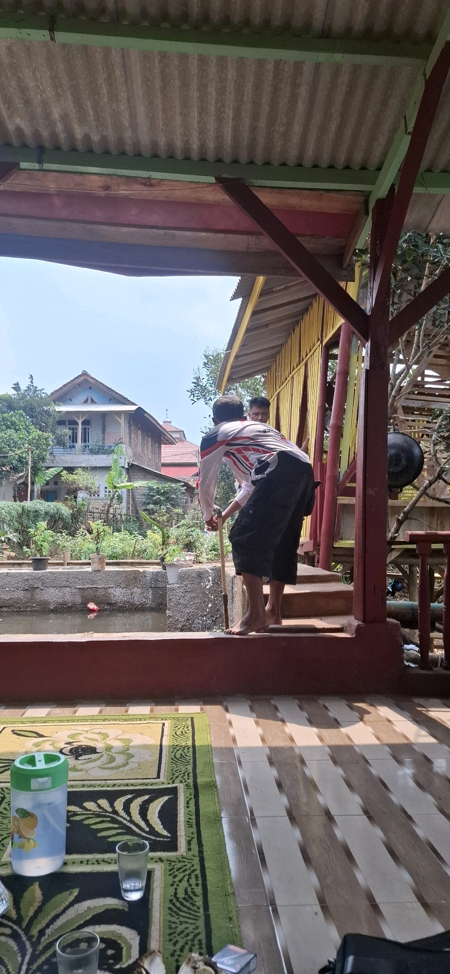 In Cilaku, Indonesia, a person wearing a white shirt with red and black accents and black shorts is seen from behind, standing on a raised platform near a pond. The individual is holding a wooden stick and appears to be interacting with the water. Another person is partially visible behind the first, looking towards the pond. The background shows houses, lush greenery, and a clear sky. In the foreground, a patterned rug is laid out on a tiled floor, with a plastic water jug, glasses, and a pack of cigarettes visible. The scene appears to be an outdoor area with a covered roof structure.