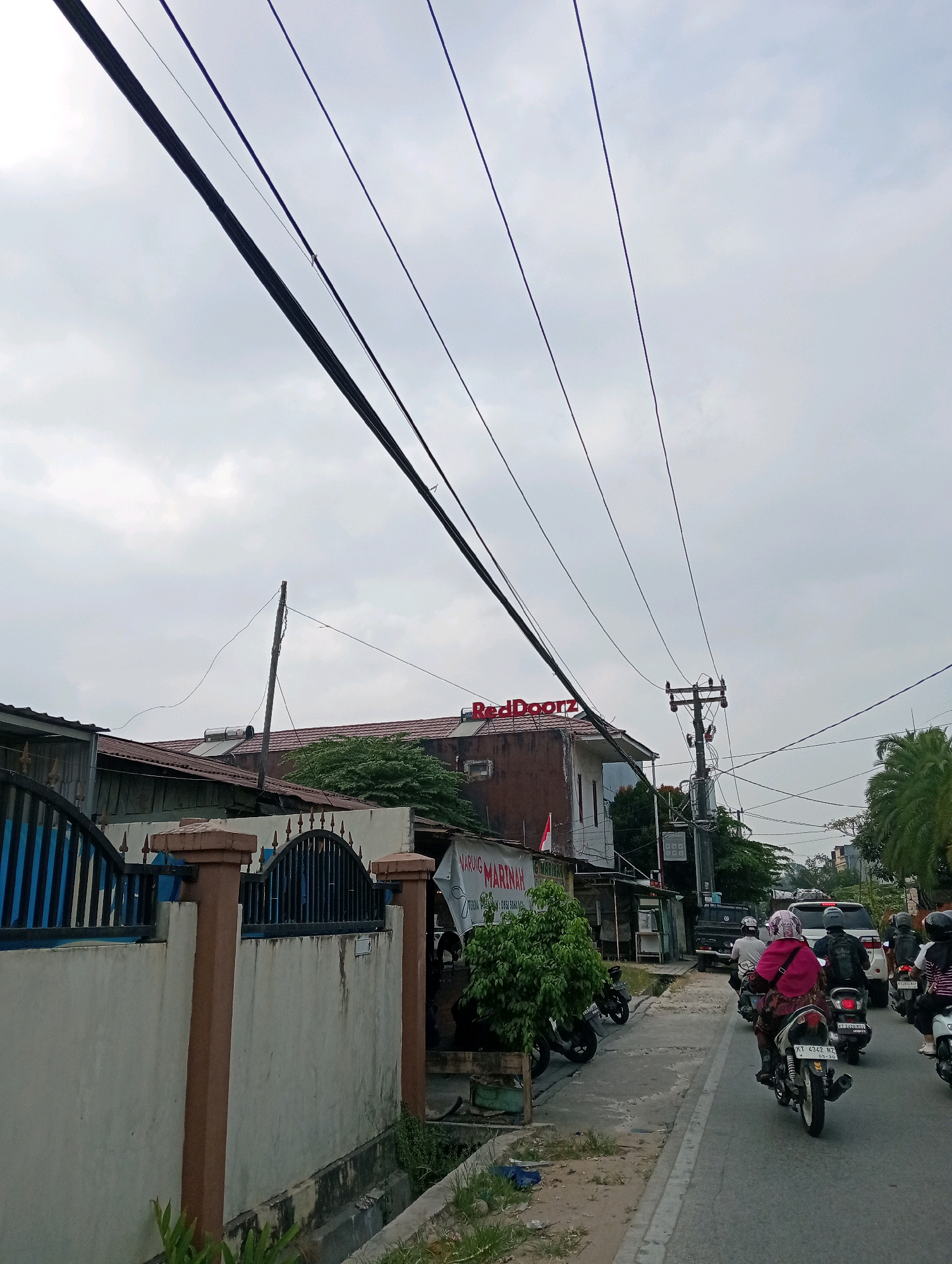 The image depicts a street scene in Samarinda, Indonesia, characterized by overcast skies and overhead power lines. In the foreground on the left, a cream-colored wall with brown pilasters and an ornate black metal fence runs along the side of the road. To the right of the wall, a paved road is visible, with several motorcycles and a white SUV in traffic. A woman wearing a pink headscarf and traditional clothing is riding a motorcycle in the foreground. Behind her, other motorcyclists and a car are also present. Buildings line the right side of the road, one of which features a red "RedDoorz" sign. A national flag is displayed on one of the buildings. Lush green trees and vegetation are interspersed throughout the scene, and a drainage ditch runs alongside the road. The overall impression is that of a moderately busy urban environment on a cloudy day.