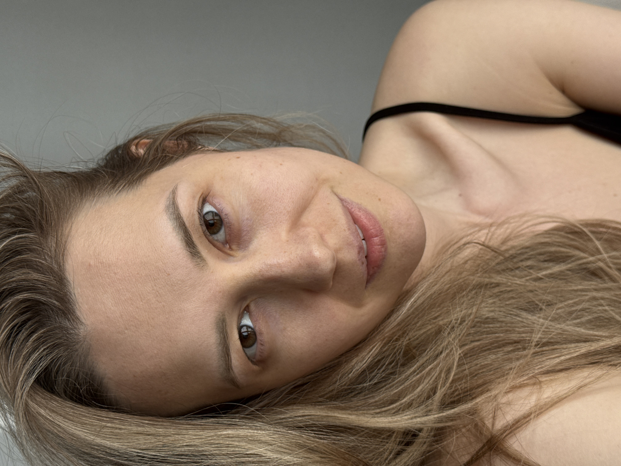 A woman with fair skin and light brown hair lies down, looking towards the camera. She has brown eyes and a slight smile. Her hair is spread out around her head and shoulders. She is wearing a black top with a thin strap. The background is a plain, light gray wall. The image appears to be taken indoors. The location of Chaikovsky, Russia, is not evident from the image.