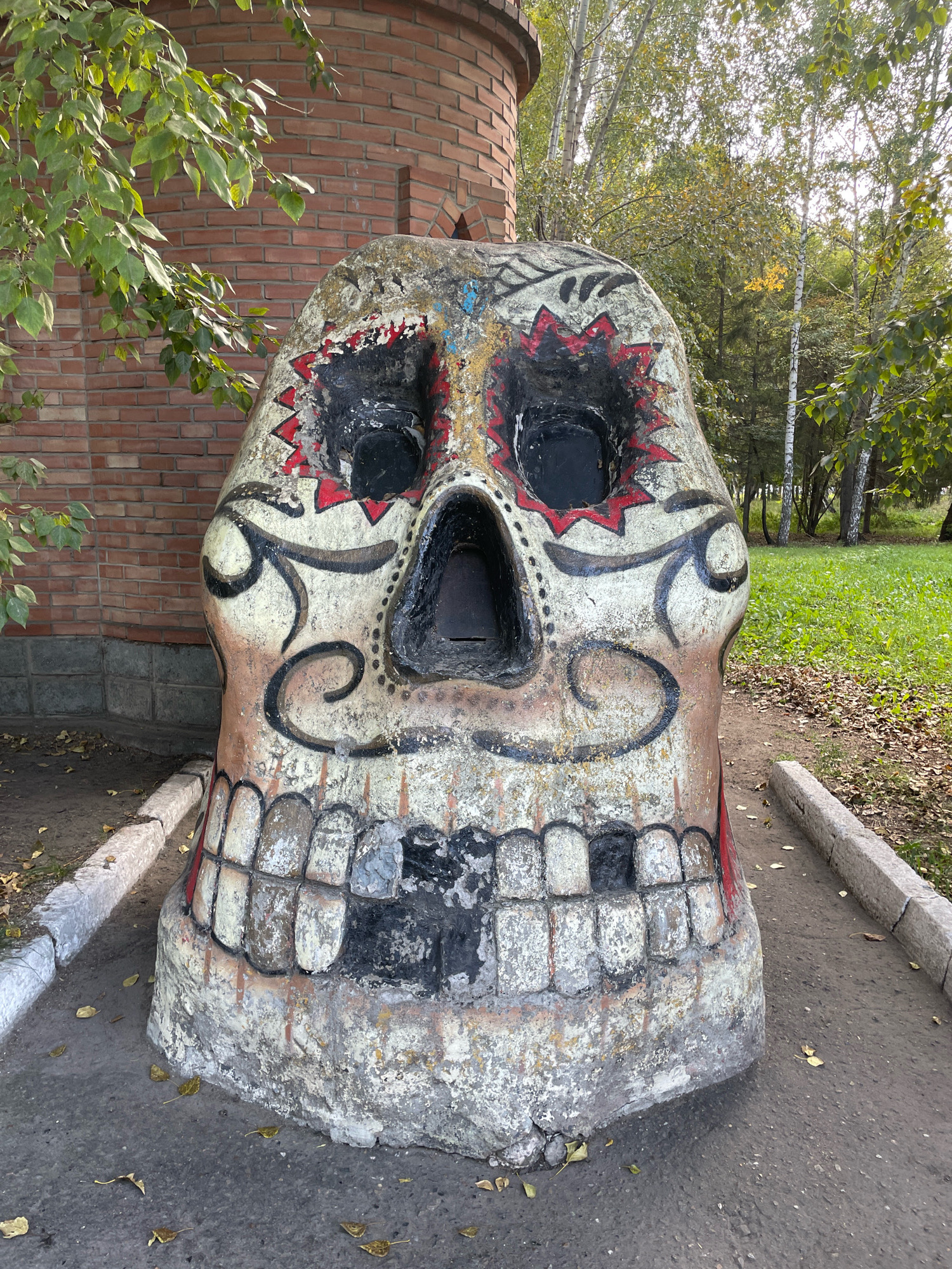 The image depicts a large, weathered, concrete skull sculpture painted with colorful designs, possibly representing a Sugar Skull from Day of the Dead celebrations. The sculpture is situated outdoors on an asphalt pathway, bordered by a concrete curb. Behind the sculpture is a brick building on the left, and a grassy area with trees and fallen leaves on the right, indicating an autumn setting. The skull itself is decorated with black, red, and yellow paint, with empty sockets for eyes and a gaping mouth. The overall condition of the sculpture suggests it has been exposed to the elements. The location is identified as the Omsk State Museum of Local History in Omsk, Russia. There are no people visible in the image.