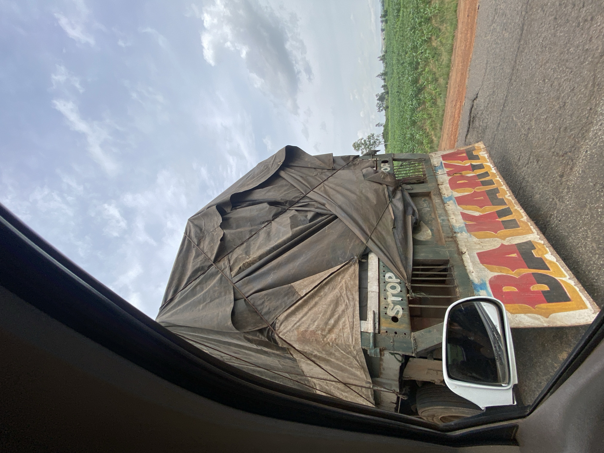 A side view of a large, covered truck on a paved road is captured through the window of a vehicle. The truck's cargo is covered by a dark, weathered tarp, secured with ropes. The truck has "STOP" stenciled in white on its side, and the word "BAKARYA" is painted in large, colorful letters on the rear. A white side-view mirror is visible in the foreground, reflecting the interior of the car and the truck. The truck is traveling through a rural area, with a field of green crops and trees visible in the background under a cloudy sky. The location is stated to be Kuru, Nigeria.