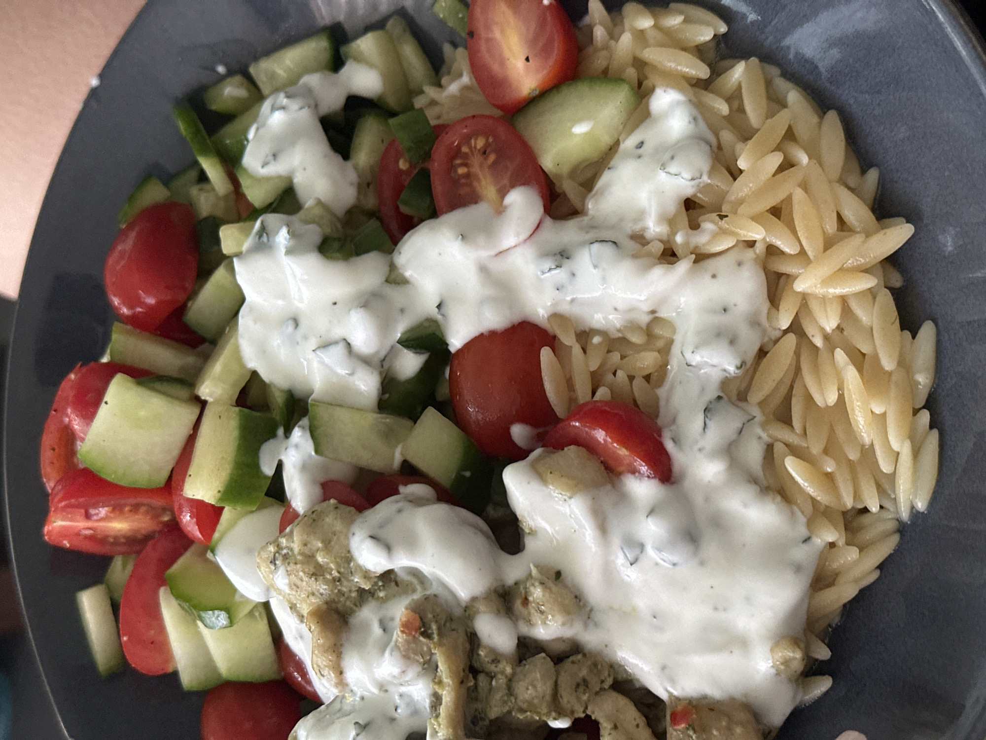 A close-up, overhead shot shows a grey bowl filled with a meal. The meal includes orzo pasta, chunks of cucumber and halved cherry tomatoes. The entire meal is topped with a white sauce. There is also what appears to be chicken pieces in the meal. The location of the image is not evident.