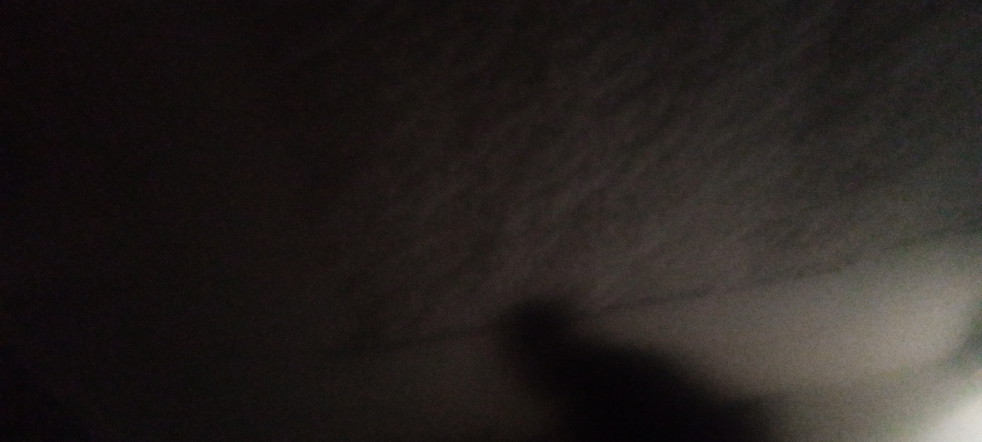 The image is a dark, blurry photograph. It appears to be taken indoors in a low-light environment. A vague human figure is visible in the lower right portion of the frame. The location is unknown.