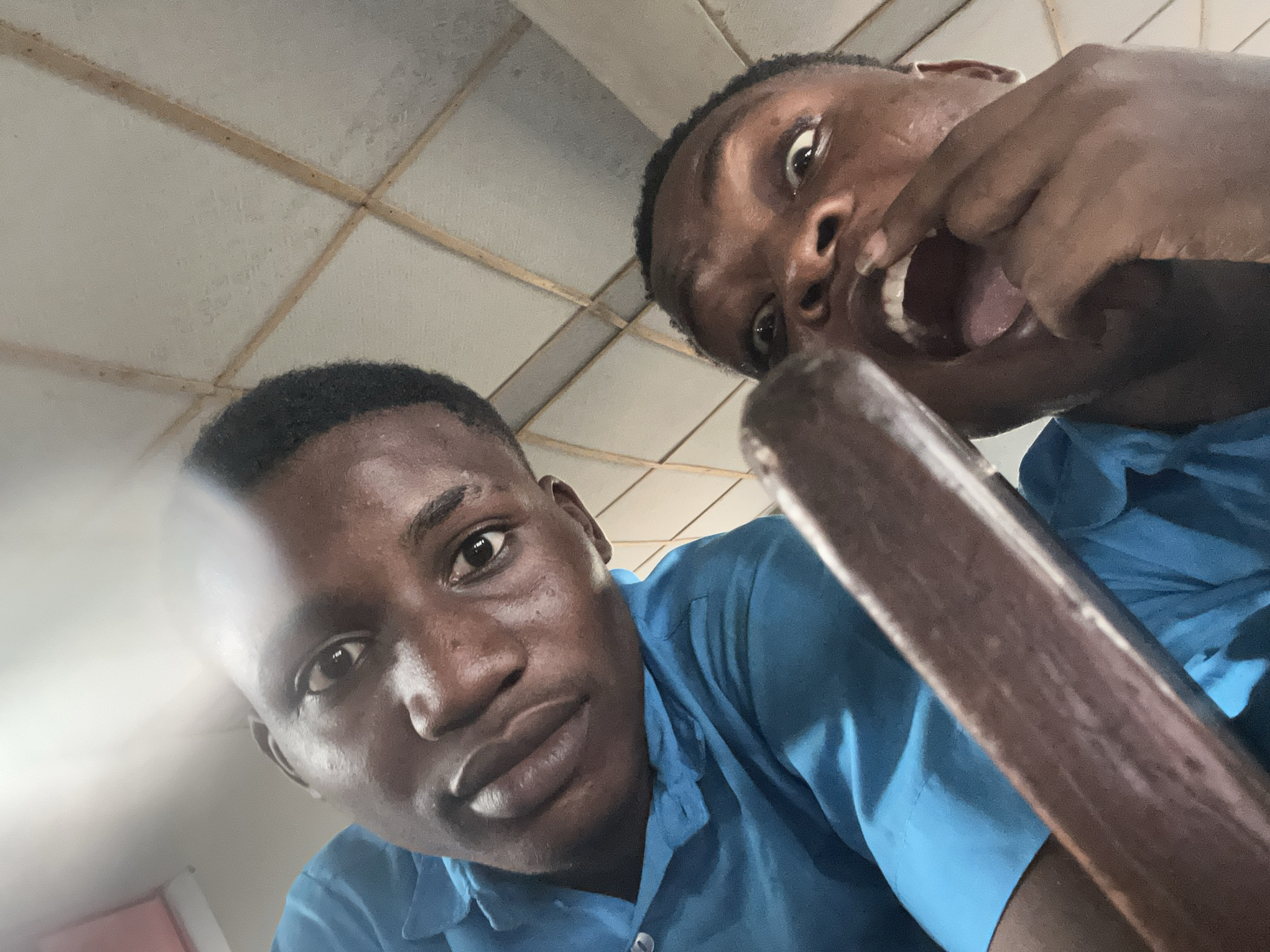 A low-angle, close-up photograph captures two young African men, appearing to be in an indoor setting. Both individuals are looking upwards towards the camera, suggesting a selfie or a shot taken from below their eye level.
The man on the left, positioned closer to the center of the frame, has dark skin, short dark hair, and a neutral to slightly curious expression, looking directly into the lens. He is wearing a light blue collared shirt.
The man on the right, situated further back and to the right, displays a more animated and humorous expression. His eyes are wide, and his mouth is open, with his right hand seemingly pulling down his lower lip or cheek to expose his teeth and tongue. He also wears a light blue collared shirt.
A dark brown, elongated object, possibly a wooden pole or handle, extends diagonally from the lower right corner, partially obscuring the man on the right.
The background reveals a light-colored ceiling composed of square tiles or panels, separated by a grid of narrow, light brown beams that show some signs of age or discoloration. The lighting is even, suggesting an indoor environment.
The playful expressions of the subjects suggest an informal and lighthearted moment, likely captured in a casual setting such as a school or community building in Ikorodu, Nigeria. There is no visible text in the image.