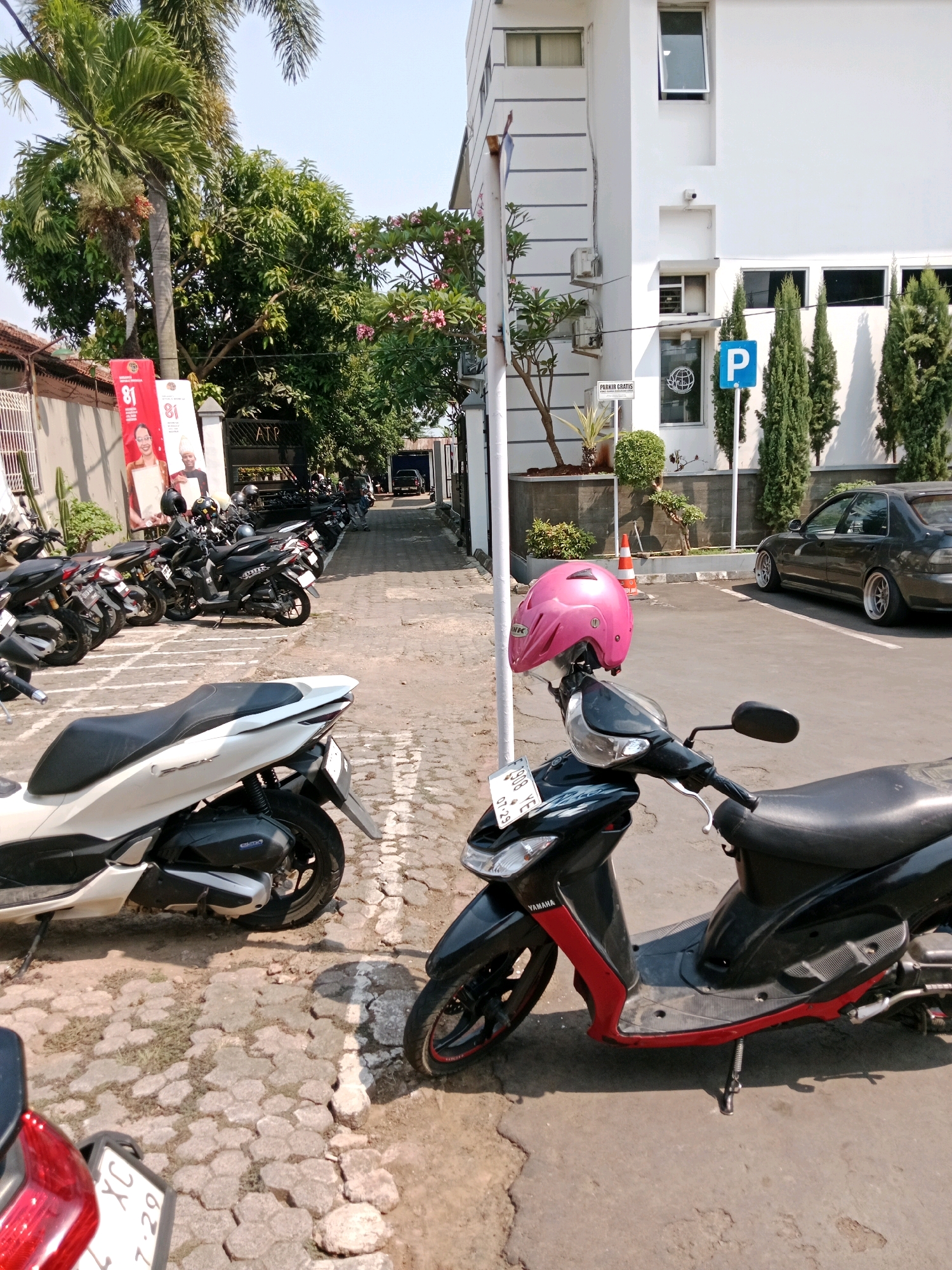 The image depicts an outdoor scene in the city of Muka, Indonesia, likely a parking area adjacent to a white building. In the foreground, two scooters are parked on a cobblestone surface. The scooter on the right is black with red accents and has a pink helmet placed on its handlebars. The scooter on the left is predominantly white. A black car is parked in the background on an asphalt surface. Several other motorcycles are visible parked in a row further down the path. Trees, including palm trees, and lush green foliage surround the area. Banners with images and text, including the number "81", are displayed along the left side of the path. A blue parking sign with a white "P" is visible on the right side of the path, near the white building. The sky is clear and bright, indicating a sunny day.