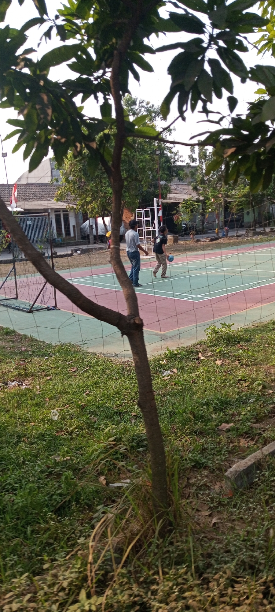 In Setu, Indonesia, on a multi-purpose outdoor court, two individuals are engaged in a game of volleyball. One person, dressed in a grey t-shirt and blue jeans, stands facing the net, appearing to be observing the game. The other individual, wearing a black t-shirt and light-colored pants, is in motion, mid-play, with a light blue volleyball in the air near them. The court itself is marked with white lines and has sections of red and green surfacing. A volleyball net stretches across the court, supported by poles. Beyond the court, a residential area is visible with houses featuring tiled roofs and visible windows. A small football goal with a net is situated on the left side of the image, near the court. The foreground is dominated by a tree with bare branches and green leaves, obscuring part of the scene. The ground is covered with grass and scattered dry leaves, with some plastic waste visible. The overall environment suggests a public recreational area.
