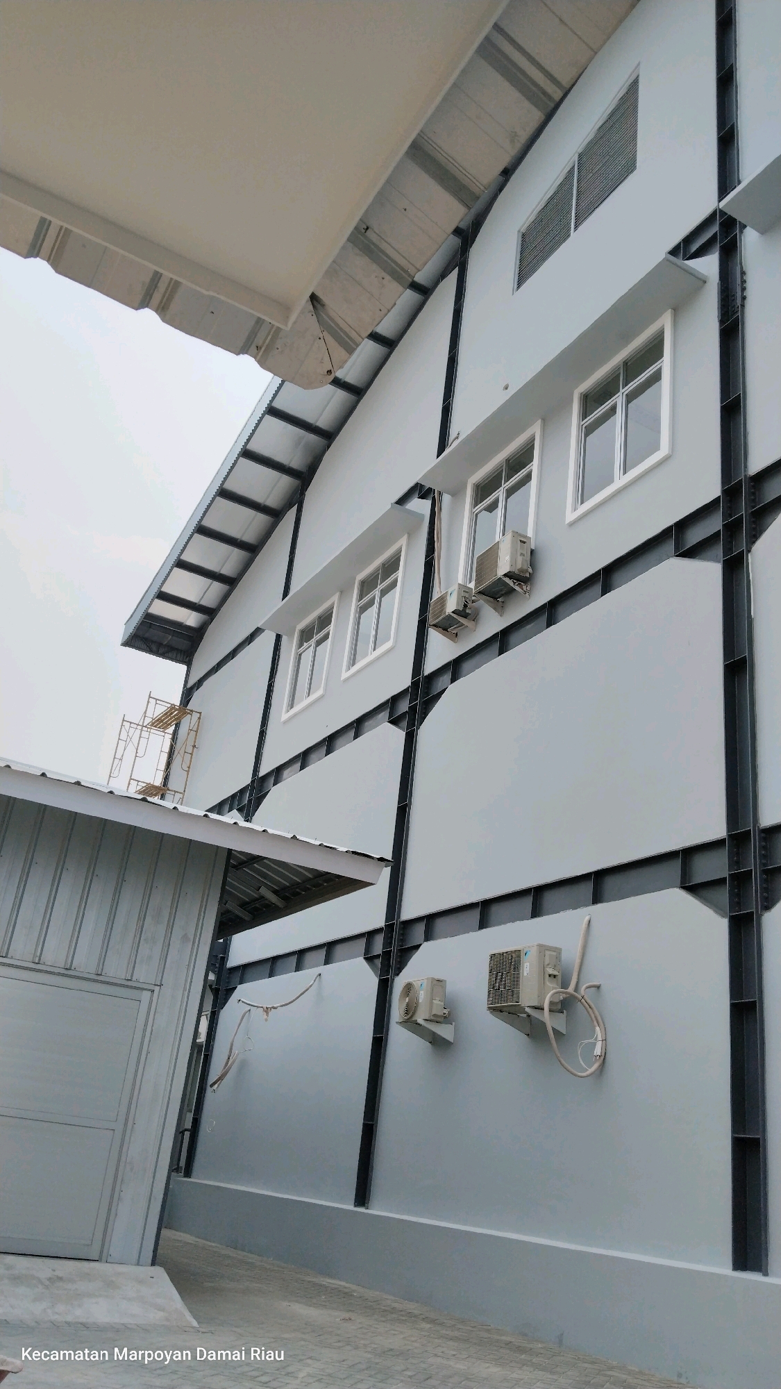 The image shows a building with a light gray exterior wall. There are several windows on the building, some with white frames and others with louvered vents. Two air conditioning units are mounted on the wall, and a clothesline is strung between two points on the building. A smaller, metal-clad structure with a corrugated roof is visible to the left of the main building. Scaffolding is erected next to this smaller structure. A sloping paved surface leads away from the smaller building. The text "Kecamatan Marpoyan Damai Riau" is visible at the bottom left of the image, indicating the location as Marpoyan Damai District, Riau, which is a district within Pekanbaru, Indonesia.