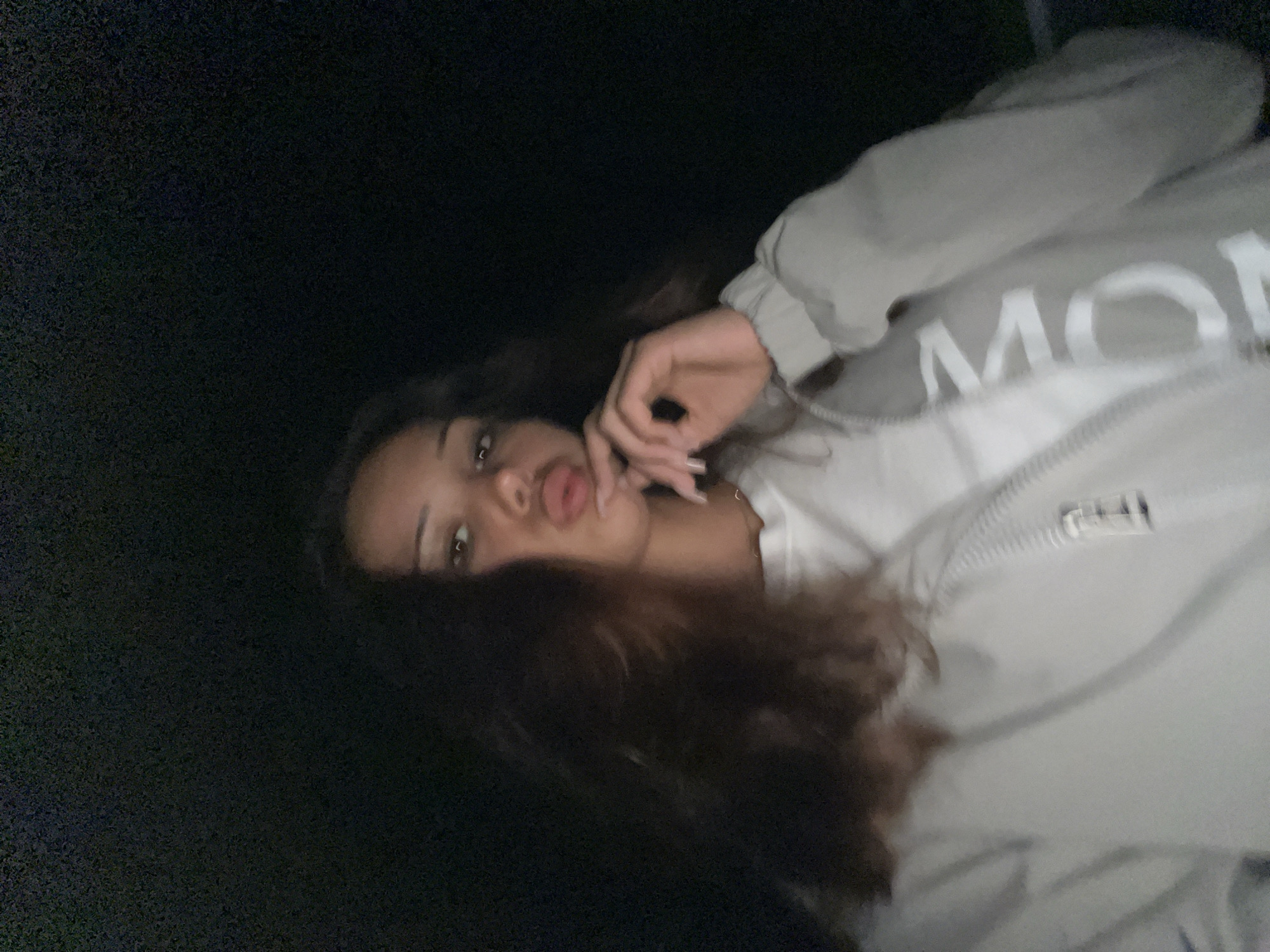 In a dimly lit outdoor setting, a young woman with long, dark, wavy hair is captured in a close-up selfie. She is wearing a light-colored hooded jacket with a zipper and a white t-shirt underneath. Her left hand is raised to her face, with her fingers lightly touching her lips. Her mouth is pursed, and her eyes are looking towards the camera. The background is dark, suggesting it is nighttime. The image does not provide any specific information about the location within Zlatograd, Bulgaria.