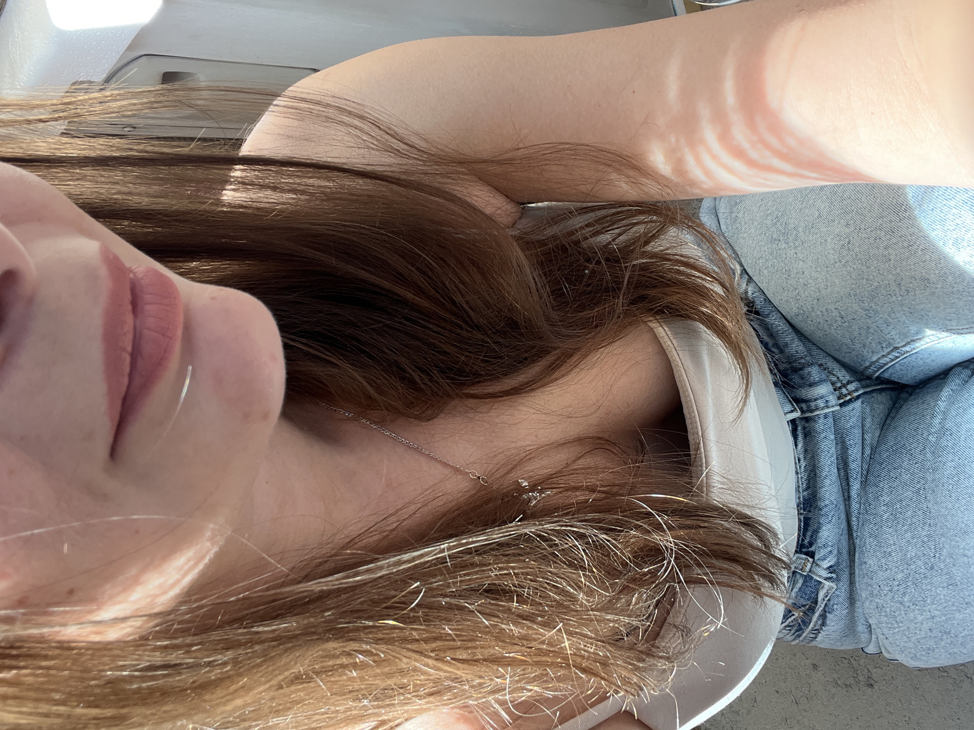 The image shows a close-up, medium-shot of a person's upper body, from the chest up. The person has long, brown hair that falls around their shoulders and chest. They are wearing a white tank top and blue jeans. A thin silver necklace with a small pendant is visible around their neck. The background is blurred and appears to be outdoors, with sunlight casting striped shadows on the person's arm. The image quality is clear, and the lighting is bright, suggesting it was taken during the day. There is no information to suggest this image is located in Tolyatti, Russia.