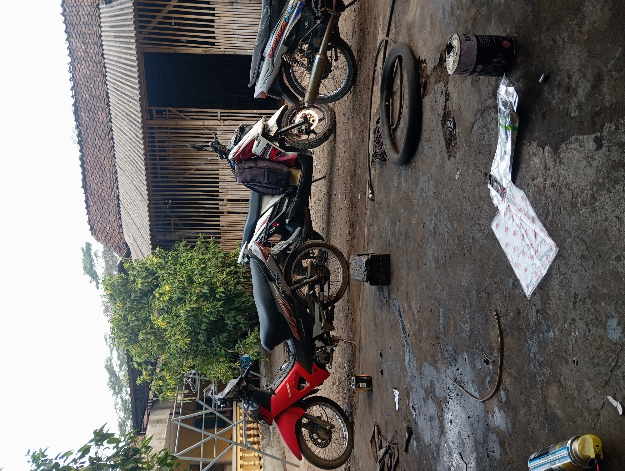 This is a medium shot, eye-level, outdoor, full-color, natural light photograph of a motorcycle repair area in Kudus, Indonesia. Three motorcycles are parked in front of a building with a corrugated metal roof and walls made of vertical bamboo poles. The ground is a mixture of dirt and concrete, with some scattered debris, including a deflated tire, a can of oil, and packaging materials. The motorcycle in the foreground is red and black with a Honda logo. The middle motorcycle is white and red with a Yamaha logo and a backpack draped over its seat. The motorcycle on the right is partially visible and appears to be silver and blue with a Yamaha logo. A tree with green foliage is visible behind the motorcycles. The scene appears to be a functional repair shop, given the presence of tools and parts.