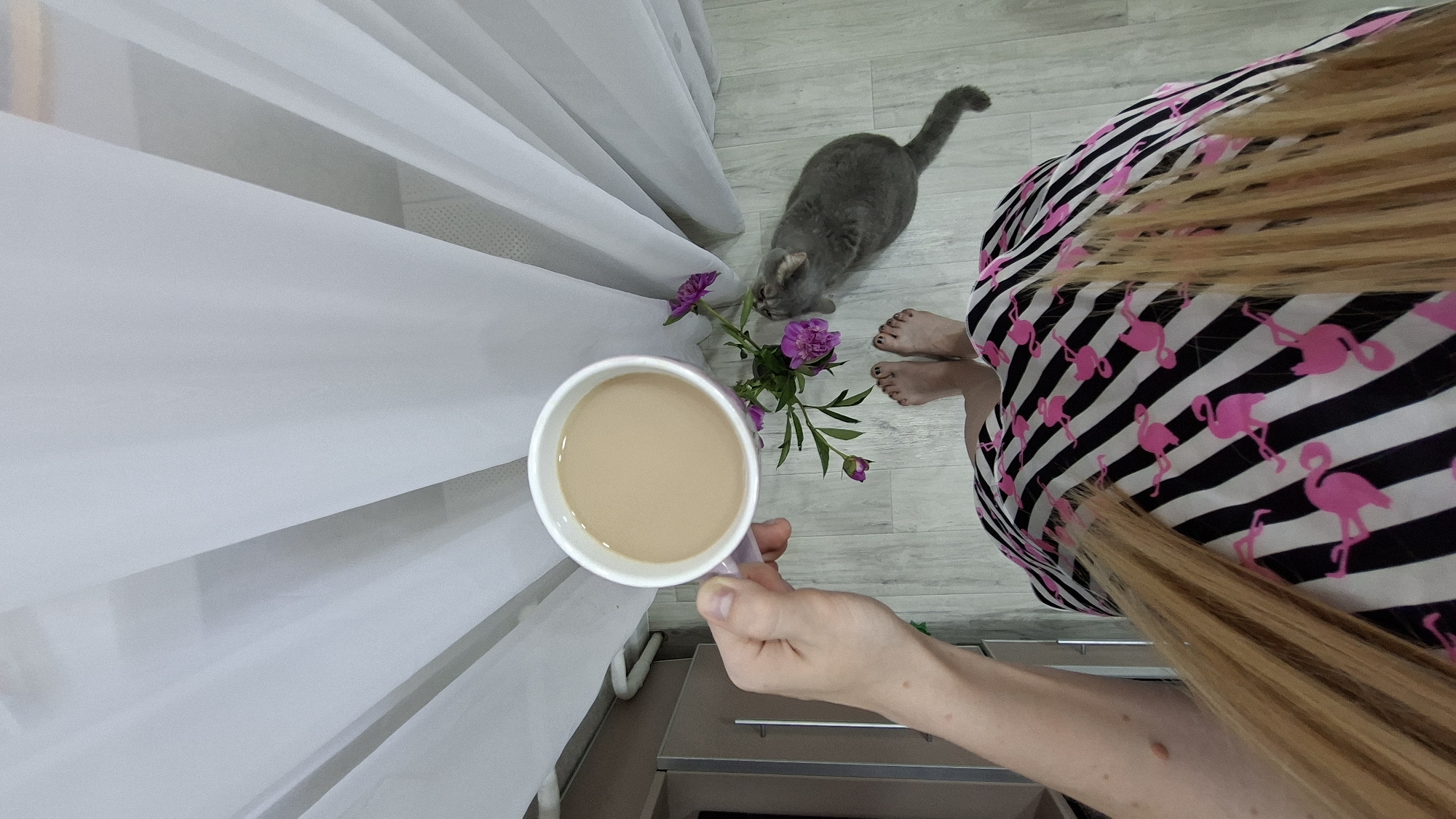A woman is holding a cup of coffee in her left hand. She is wearing a black and white striped skirt with pink flamingo patterns. Her bare feet are visible on a wooden floor. A gray cat is standing next to a small vase of purple flowers, looking at the flowers. The scene is indoors, with sheer white curtains in the background. The floor appears to be light wood laminate.