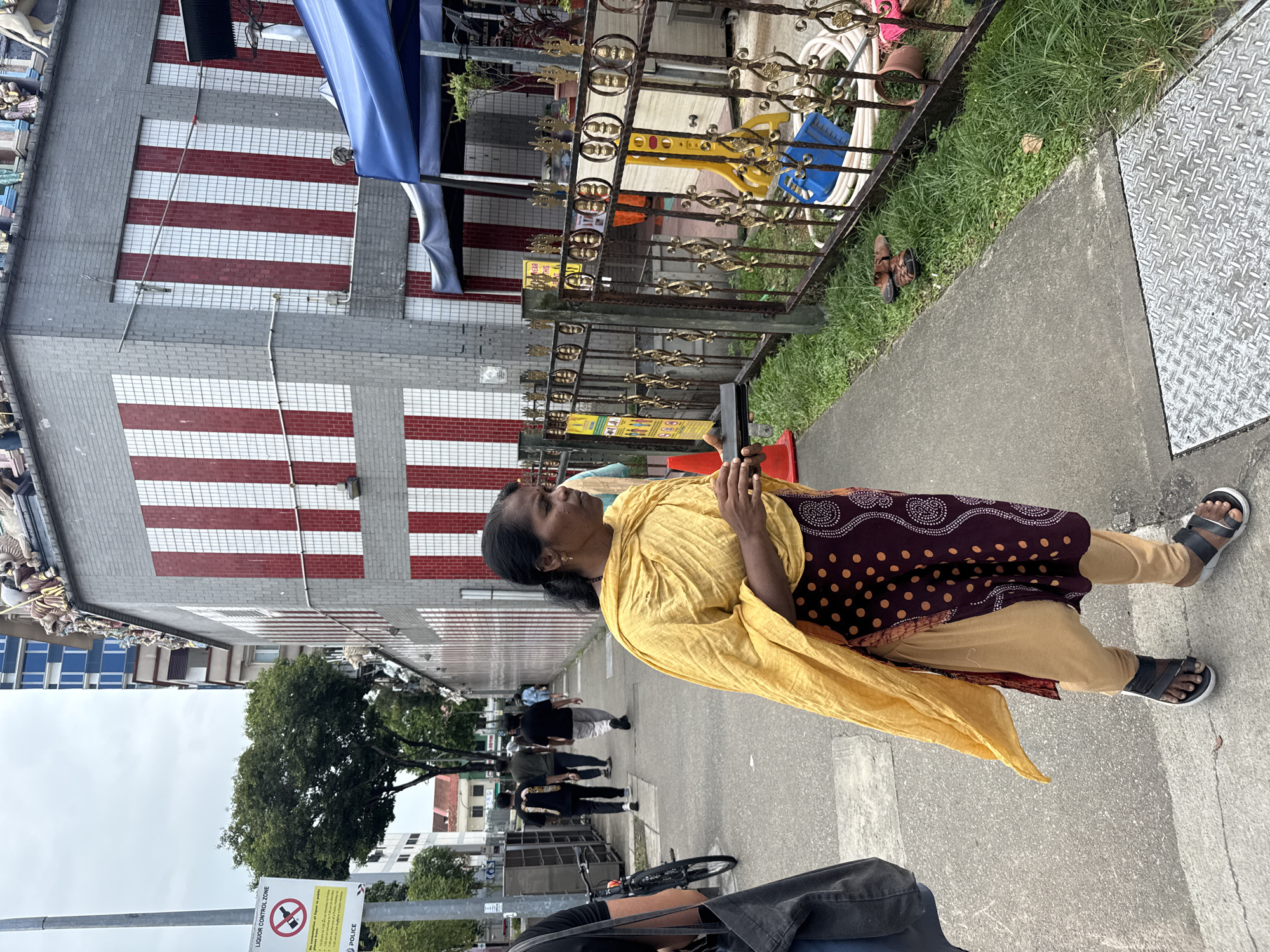 In a street scene in Singapore, a woman with dark skin and hair tied back walks on a sidewalk. She wears a yellow shawl over a dark, patterned garment and light-colored pants, with sandals on her feet. She holds a small, dark object in her hands. Behind her, several men walk away from the camera along the same sidewalk. To the right, a metal fence with ornate gold designs encloses an area with grass and some stored items, including a blue dustpan and a hose. A building with a facade of white and red vertical stripes is visible behind the fence. The building's roof is adorned with decorative figures. On the left side of the image, a sign with red lettering indicates a "LIQUOR CONTROL ZONE." In the background, a tall blue building can be seen. The sky is overcast.