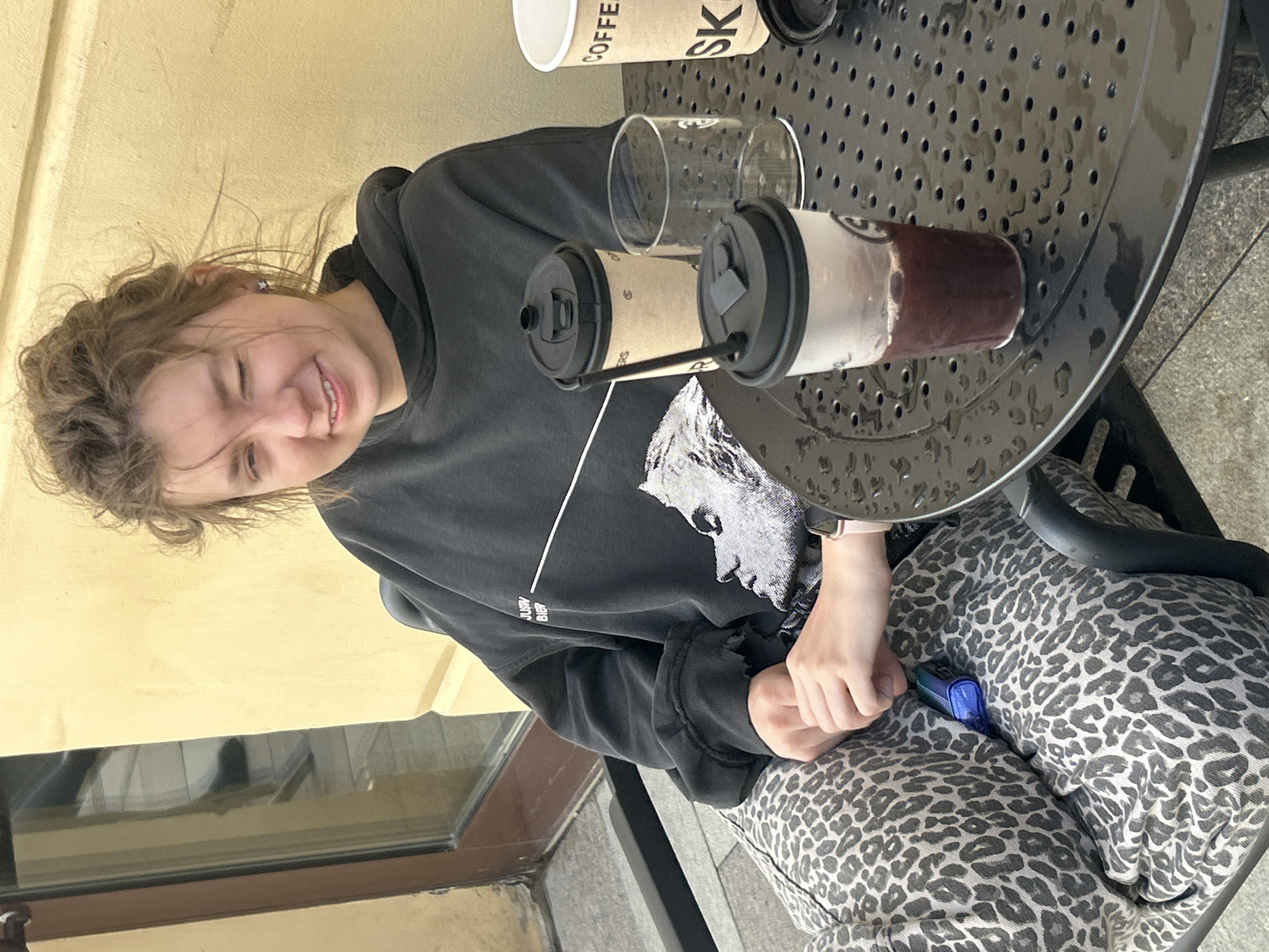 In an outdoor setting, a young person sits at a round, black metal table with a perforated surface, likely at a cafe or restaurant. The person is wearing a dark grey hoodie with a graphic print of a face and some text on the front. They are also wearing patterned pants with a leopard-like print. In front of them on the table are two paper coffee cups with lids and straws, and two clear glasses. One of the coffee cups appears to contain a dark beverage. The person is smiling and winking, suggesting a lighthearted or playful moment.
The table is wet, possibly indicating recent rain or dew, and there are some reflections on its surface. The background features a yellow wall and a glimpse of a window or glass door. The lighting suggests it is daytime, and the overall scene appears casual and relaxed. A small, blue object, possibly a vape or electronic device, is tucked into the person's pants. The image is taken from a slightly high angle, looking down at the person and the table. The hair of the person is messy and windswept.