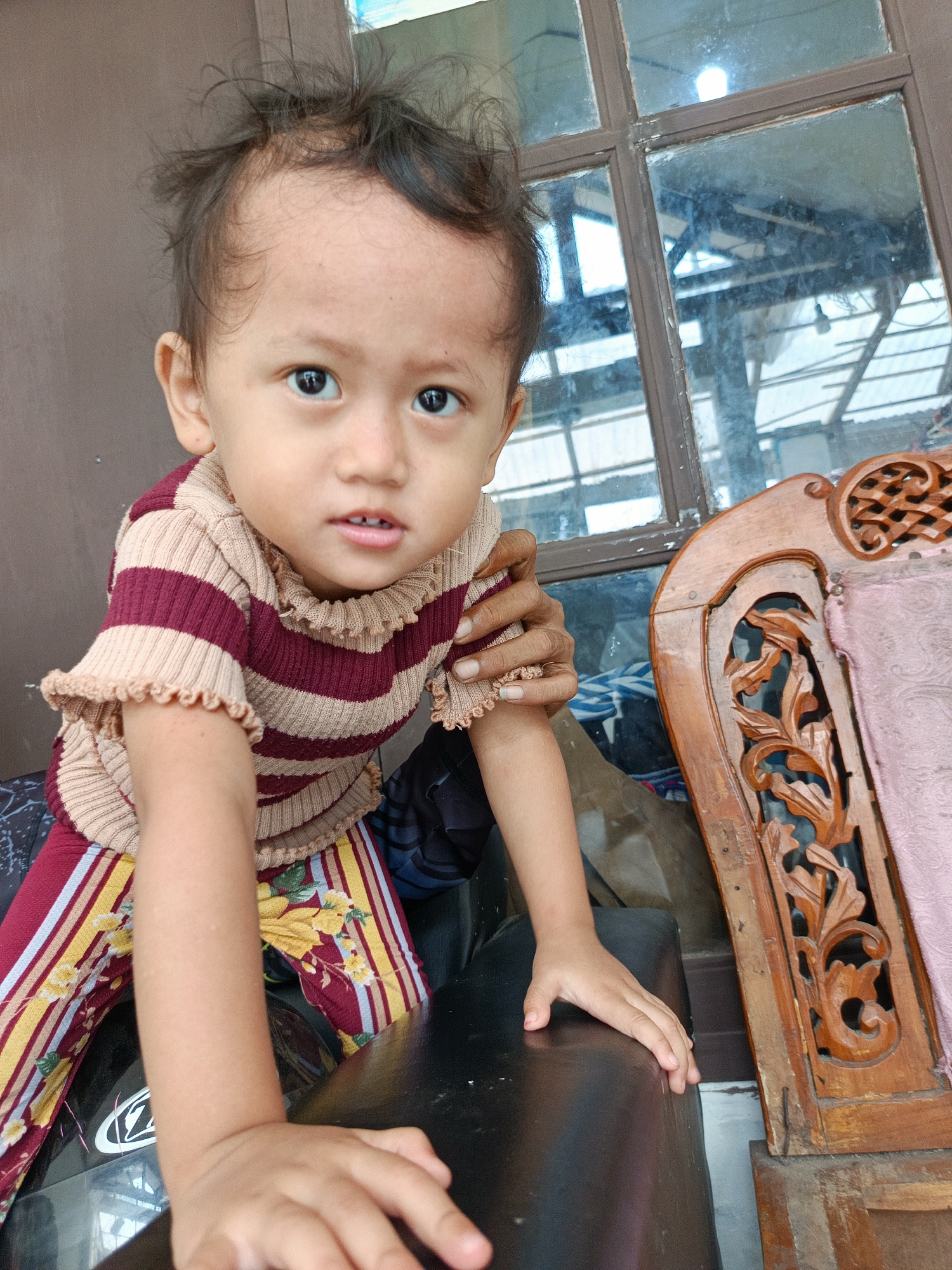A young child, appearing to be of Southeast Asian descent, is positioned in the foreground of the image. The child has dark, somewhat disheveled hair and is looking directly at the camera with wide, dark eyes. They are wearing a striped shirt in shades of burgundy and beige, with ruffled sleeves, and colorful striped pants featuring yellow floral patterns on a burgundy background. The child's hands are visible; one hand is resting on a dark, textured surface, and the other is being held by an adult, whose arm is visible from the shoulder down, wearing a ring on one finger.
The background reveals a weathered window with multiple panes, reflecting an outdoor environment. To the right of the child is a portion of an ornate wooden chair with intricate carvings and a pink upholstered seat. The overall setting appears to be an outdoor or semi-outdoor area, possibly a porch or patio, with a sense of lived-in clutter. The location is identified as Margajaya, Indonesia.
