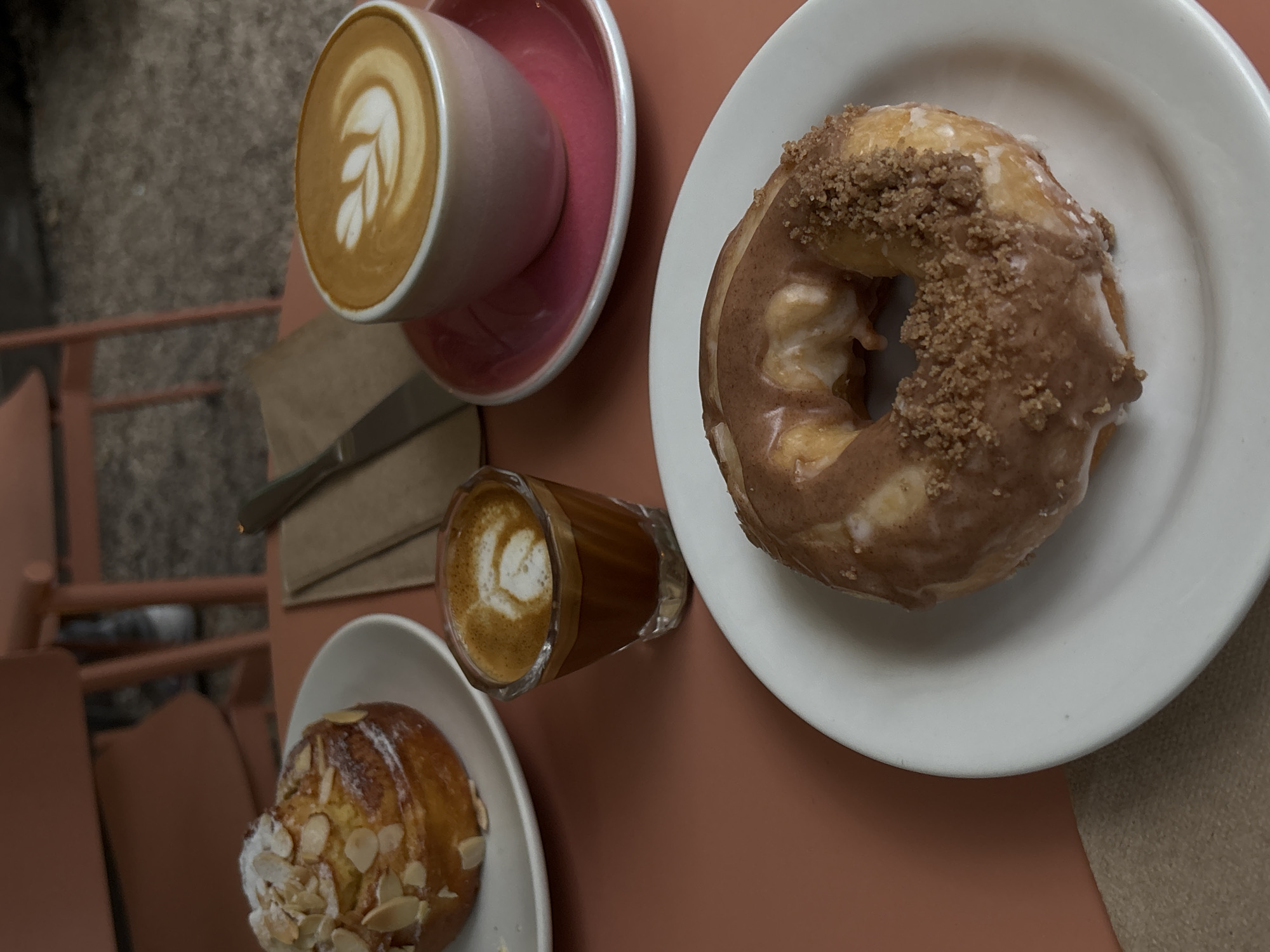 The scene presents a top-down view of a table set for coffee and pastries, presumably in a café. At the top left, a pink cup with a latte sits on a matching saucer. A knife rests beside it on a folded brown paper napkin. Adjacent is a glass containing coffee with latte art. In the top right quadrant, a frosted donut is presented on a white plate. Towards the bottom left, a pastry topped with almonds is on a similar white plate. The table itself is a solid, muted reddish-brown color. Parts of metal chairs are visible on the left, suggesting outdoor seating. No individuals are discernible.
