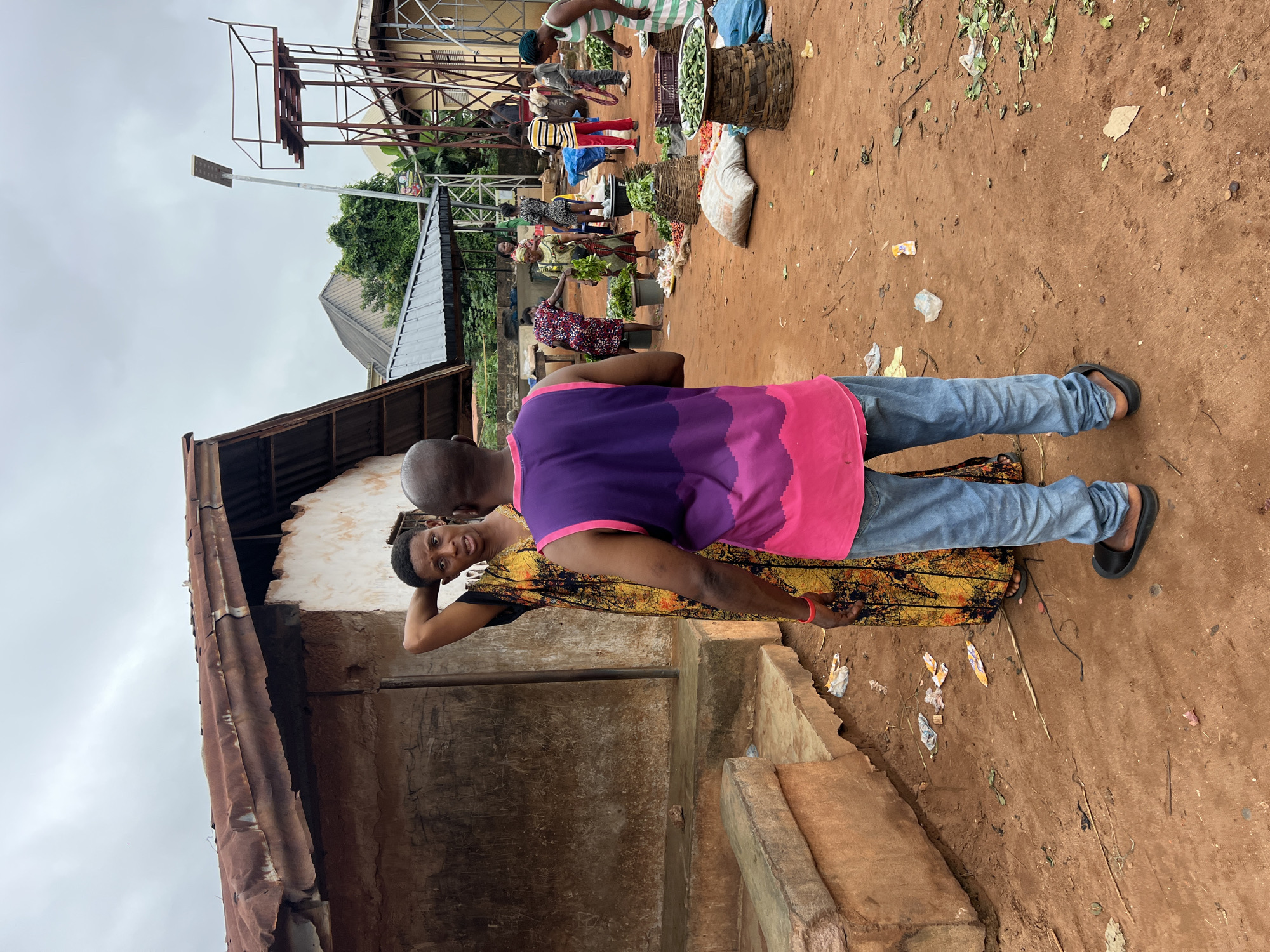In Nkpor, Nigeria, at an outdoor market on a cloudy day, a man wearing a purple and pink tank top and blue jeans stands facing away from the camera, engaged in conversation with a woman. The woman, dressed in a vibrant yellow and black patterned dress, stands to his left with her left hand on her head. Behind them, other market-goers are seen amidst stalls of produce, including baskets of vegetables and what appear to be sacks of goods. A rustic, weathered building with a corrugated metal roof is visible to the left, and a tall metal structure with a water tank and a solar-powered light stands in the background. The ground is predominantly dirt, with scattered debris and fallen leaves.