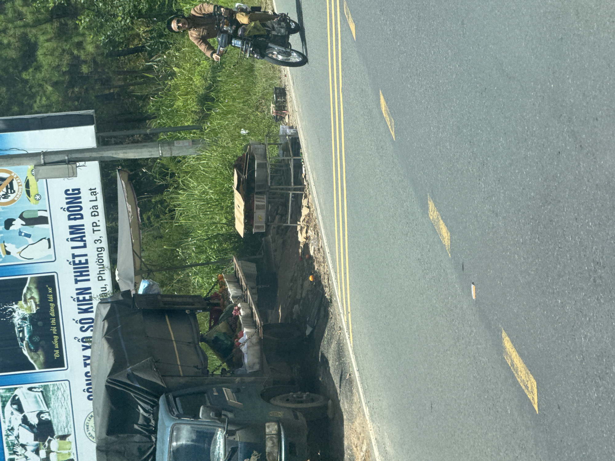 The image depicts a roadside scene in Thôn 6, Vietnam. In the foreground, a grey asphalt road with yellow lane markings is visible. To the left of the road, a dark green truck is parked, its bed covered by a dark tarp. The truck appears to be loaded with various items, including baskets of what seem to be fruits and vegetables. Next to the truck, a makeshift stand is set up with more baskets and some stacked cardboard boxes. In the middle ground, a person wearing a light brown jacket and sunglasses is riding a motorcycle. Behind the roadside stall and the motorcycle, dense green vegetation and trees form the background, suggesting a rural or semi-rural environment on the outskirts of a city. A billboard with text in Vietnamese is partially visible above the roadside stall.