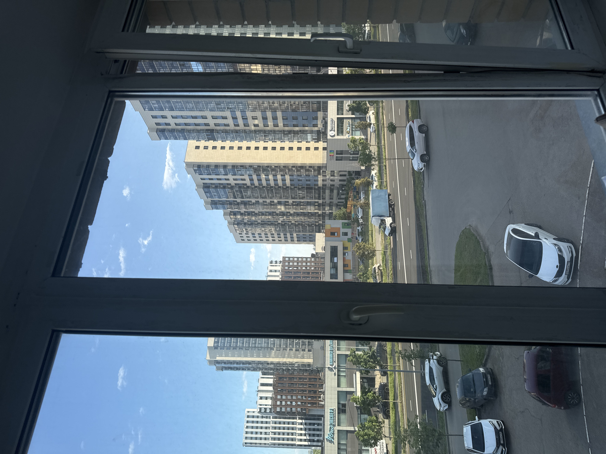 This image captures a view from a window overlooking a street and several modern apartment buildings in Kazan, Russia. The sky is blue with scattered white clouds. The buildings are tall and contemporary, with many windows, reflecting the daylight. In the foreground, through the window frame, a street is visible with cars parked and driving. Some of the cars are white, black, and maroon. Trees line the street. There are no people clearly visible in the image. The scene suggests a residential and commercial area.