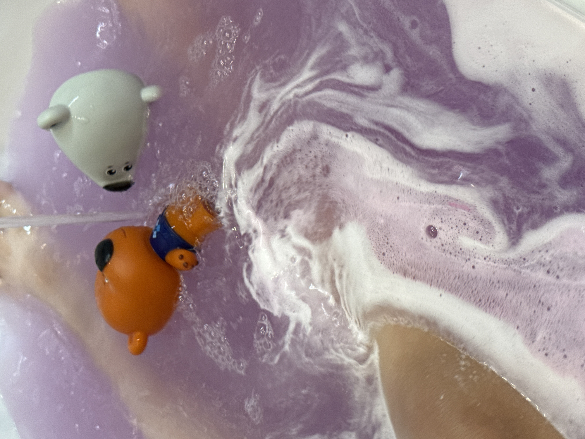 A top-down, close-up shot shows two bath toys submerged in purple water with white foamy swirls. On the left, a small, white, polar bear-shaped toy with black eyes and mouth floats. To its right and slightly lower, an orange, pig-shaped toy wearing a blue collar is partially submerged, with its snout pointing forward and its tail curled. The water is agitated, creating foamy patterns. The scene appears to be a bathtub or a similar water-filled container. There is no visible text, and no specific time of day, weather, or location cues are apparent beyond the general setting of a bath. The mood is playful and relaxed.