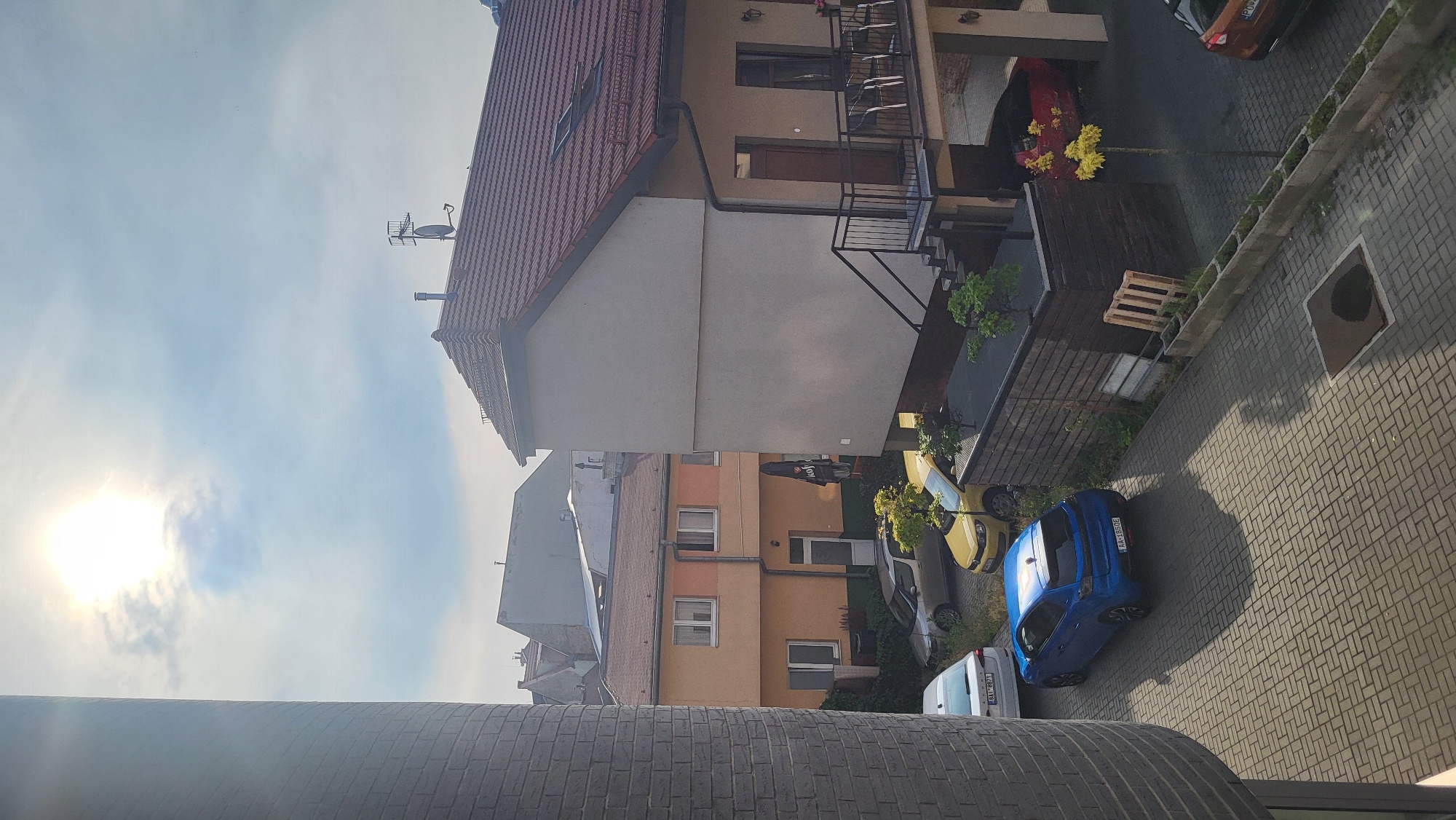 A vertical, eye-level, outdoor photograph captures a sunny day in a residential area. In the foreground, a circular, grey brick structure occupies the left side of the frame. Beyond this, a paved courtyard of grey interlocking bricks leads to several parked cars and buildings. A bright blue hatchback is parked centrally on the paving. To its left, a white sedan is parked. Further back, a silver hatchback and a yellow hatchback are visible.
The background features a two-story building with an orange facade and white-framed windows. Adjacent to this, a larger building with a grey facade and a red tiled roof dominates the right side of the image. This building has a balcony with a black metal railing and stairs leading up to it. A red car is parked in the covered garage area beneath this building.
The sky is bright blue with scattered white clouds, and the sun is high and shining, casting distinct shadows. The environment appears to be a typical urban or suburban residential setting.