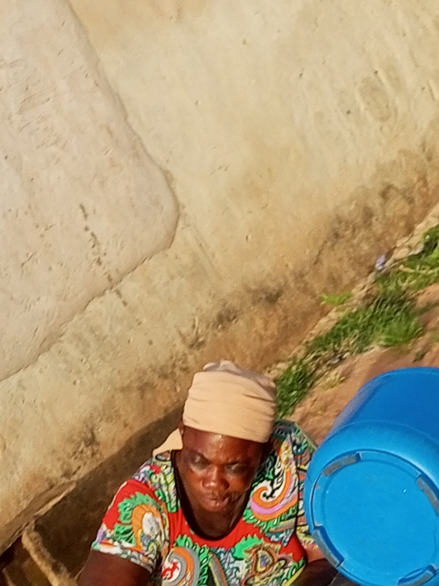 An overhead, high-angle medium shot shows a Black woman from the chest up, viewed from above, as she sits on the ground with a blue plastic bucket to her right. She wears a light tan headwrap and a colorful short-sleeved shirt with a repeating abstract pattern in red, green, orange, blue, and black. Her head is bowed, and her eyes are closed as if she is concentrating or resting. She appears to be in an outdoor, possibly rural setting. The ground is made up of dry, dusty earth with patches of sparse green grass growing along a slanted embankment on the right side of the frame. A textured, pale tan wall or structure takes up the left two-thirds of the background. The scene is lit by bright sunlight, casting subtle shadows.