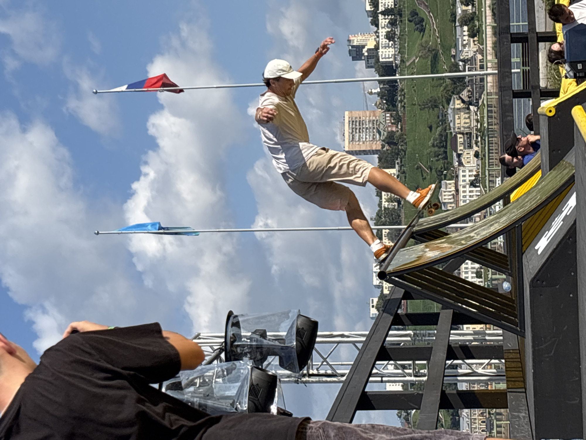 In Nizhny Novgorod, Russia, under a partly cloudy blue sky, a male skateboarder in a white t-shirt, beige shorts, and a white cap is captured mid-air, performing a trick on a ramp. He is balanced on his skateboard, with his arms outstretched for stability. The ramp is made of wood with black metal railings. To the left, a person in a black t-shirt is visible, holding a camera and looking towards the skateboarder. In the background, a cityscape with multi-story buildings and a green, hilly landscape is visible. Two flags, one blue and one of the Russian flag, are flying on flagpoles in the distance. The scene appears to be an outdoor event or competition.