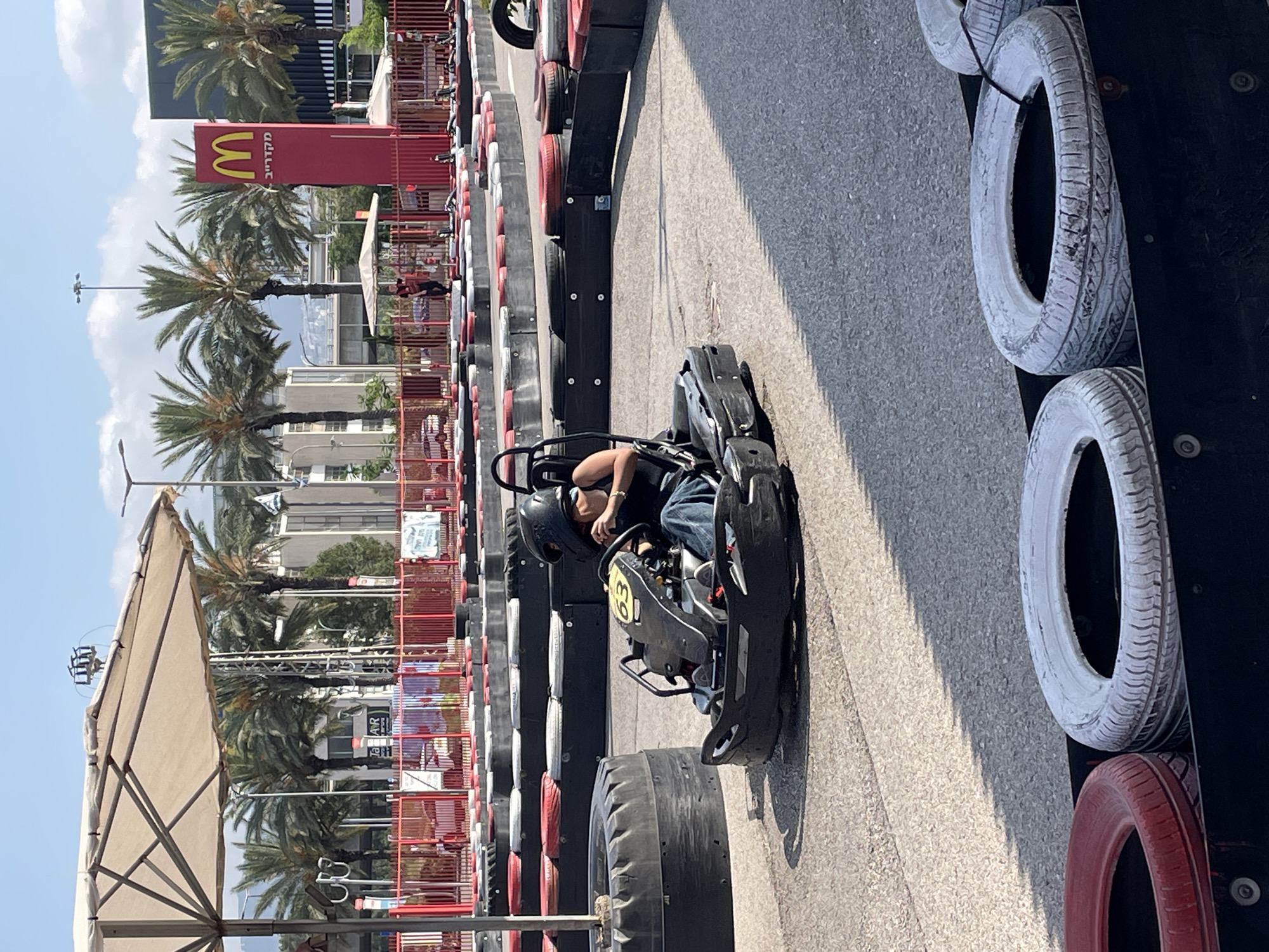 A person wearing a black helmet and dark clothing is driving a go-kart with the number 63 on it, around a go-kart track. The track is made of asphalt and is lined with tires, some painted red and white. In the background, there is a McDonald's restaurant with its iconic red sign. Palm trees are visible in the distance, and the sky is blue with some clouds. The scene appears to be outdoors on a sunny day. The location is identified as Haifa, Israel.