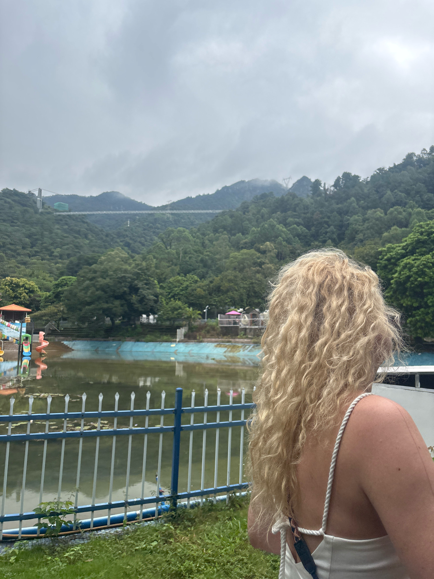 In the city of Sanshui District, China, a blonde woman stands with her back to the camera, her curly hair cascading down her shoulders. She is wearing a white tank top with rope straps. In front of her, a body of water, likely a drained or disused swimming pool, reflects the overcast sky. Beyond the pool, a children's play area with colorful slides is visible on the left. In the background, a lush, green mountainous landscape is dotted with trees. A long suspension bridge stretches across the mountains in the distance. The weather appears to be cloudy.