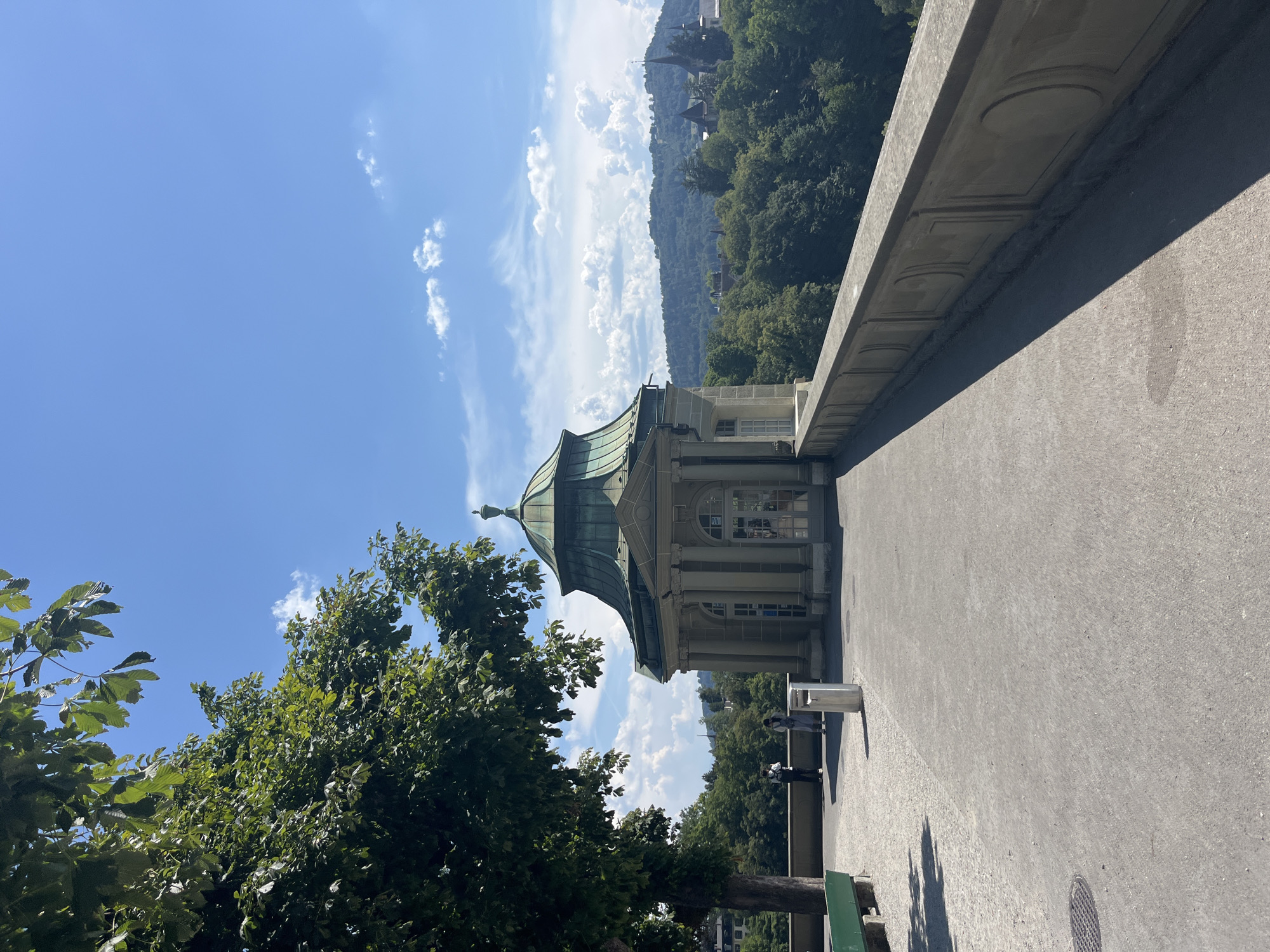 This image depicts a view from the Rose Garden (Rosengarten) in Bern, Switzerland. In the foreground, a paved walkway leads towards a classical-style pavilion with a green domed roof. The pavilion features columns and arched windows. To the left, a large tree with lush green foliage extends into the frame. A green park bench is visible on the far left. Two individuals, one wearing dark clothing and a backpack, and another in lighter clothing, stand near a trash receptacle. A stone balustrade runs along the right side of the walkway, overlooking a densely forested valley. In the background, rolling hills covered in trees rise, with several buildings, including spires, visible amidst the greenery. The sky is a clear blue with scattered white clouds. The scene is bathed in sunlight, casting shadows on the walkway.