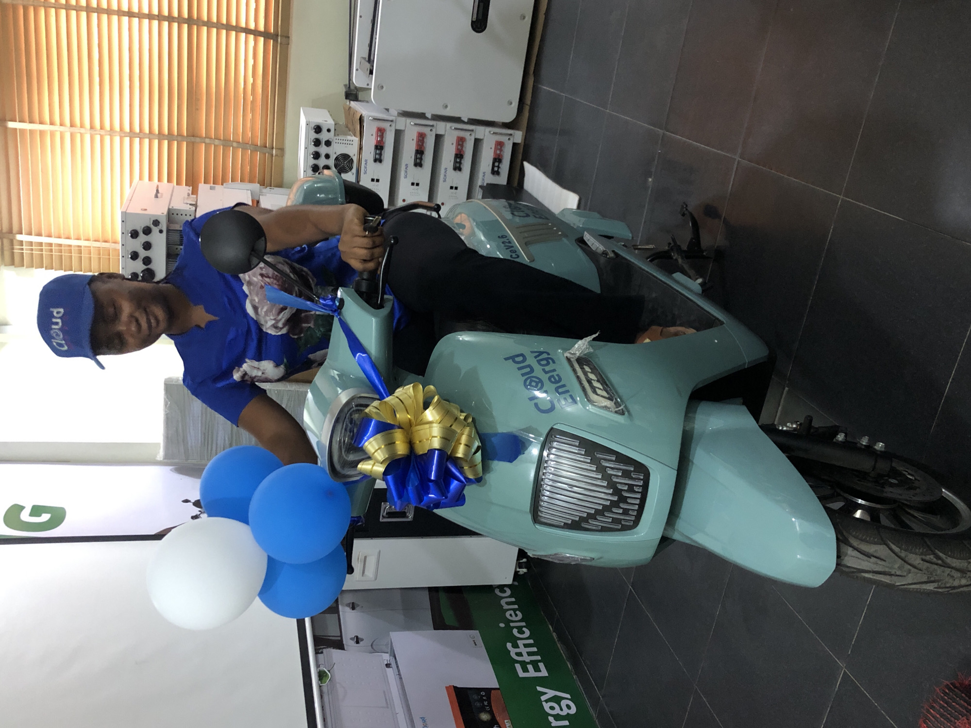 A woman wearing a blue shirt and a blue cap sits on a light teal colored scooter in what appears to be a showroom or storage facility. The scooter has a large blue and gold bow tied to the front. Three balloons, one white and two blue, are attached to the handlebars. In the background, there are stacks of white electronic equipment with visible wiring and labels. The floor is tiled in a dark color, and there are vertical blinds on a window behind the woman. A banner on the left side of the image reads "Energy Efficiency". The scene is well-lit.