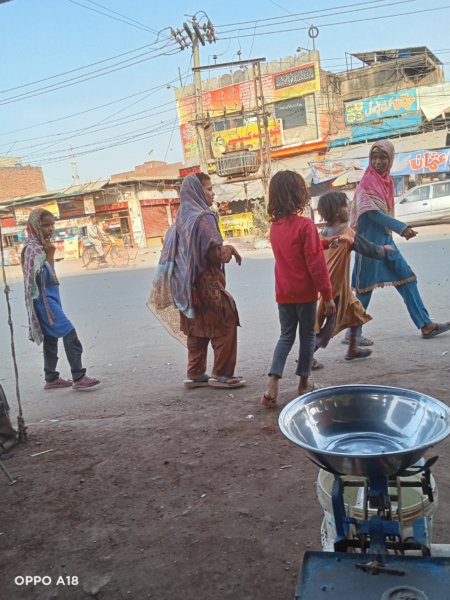 A bustling street scene unfolds under a bright, clear blue sky, likely in a South Asian locale. In the foreground, a large, shiny metallic bowl rests on a blue weighing scale, which is itself placed on a white bucket, suggesting a vendor's setup or market area. The ground is dusty and unpaved in this section.
A group of five people, primarily women and children, walk from left to right across the mid-ground. From left to right: a young girl or woman in a light blue outfit and a patterned headscarf (light green, pink, white) looks slightly to her right, wearing black pants and pink sandals. Next, a woman dressed in a brown and purple patterned outfit with a matching head covering, wearing brown sandals. Following her is a child in a red long-sleeved top and dark pants, with dark hair and black sandals. Another child in a light brown tunic walks between the child in red and the woman on the far right. The person on the far right is a woman in a blue shalwar kameez and a pink headscarf, looking back over her left shoulder, wearing dark sandals.
The background reveals a vibrant street lined with multi-story buildings housing numerous shops. Colorful signs, some bearing Arabic or Urdu script, adorn the facades. Visible text includes "BAR B," "24" (possibly indicating hours of operation), "ماشاء الله لا قوة الابالله" (Masha'Allah la quwwata illa billah - "What God wills, there is no power except with God"), "والله خير الورقي" (Wallahu Khairur-Raziqin - "And Allah is the best of providers"), "مان کر الیاس عملی حمیت کی بابان" and "عثمان" (Usman). Many utility wires crisscross overhead, connected to prominent utility poles. A white car is partially visible on the right side of the road, and a man on a bicycle can be seen further down the street on the left. In the distant background on the left, a tall minaret of a mosque stands against the sky. The overall scene depicts everyday activity in a lively urban or semi-urban environment. The bottom left corner of the image has an "OPPO A18" watermark.
