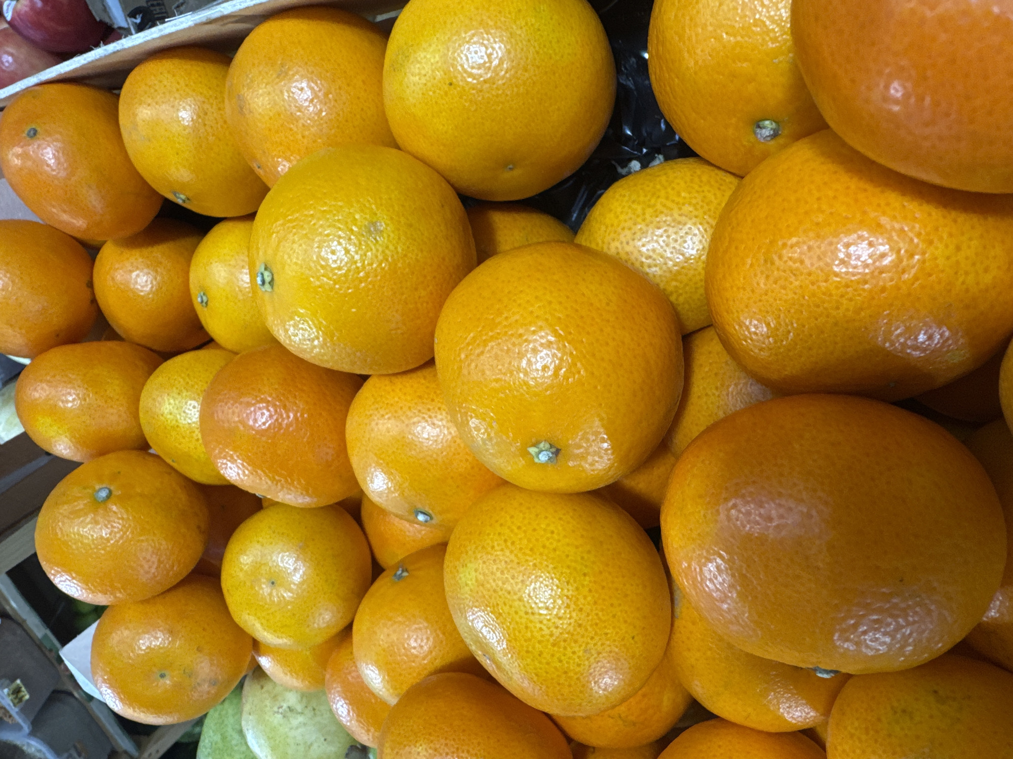 A close-up, eye-level shot captures a large pile of ripe, bright orange tangerines. The fruits are densely packed together, filling the frame. Their textured skins, characteristic of tangerines, are clearly visible, reflecting light and highlighting their freshness. The arrangement suggests a market stall or a display in a grocery store. The background is blurred, but hints of wooden crates and other produce can be discerned. The image is well-lit, emphasizing the vibrant color of the tangerines. The scene is devoid of any human presence or activity, focusing solely on the fruit.